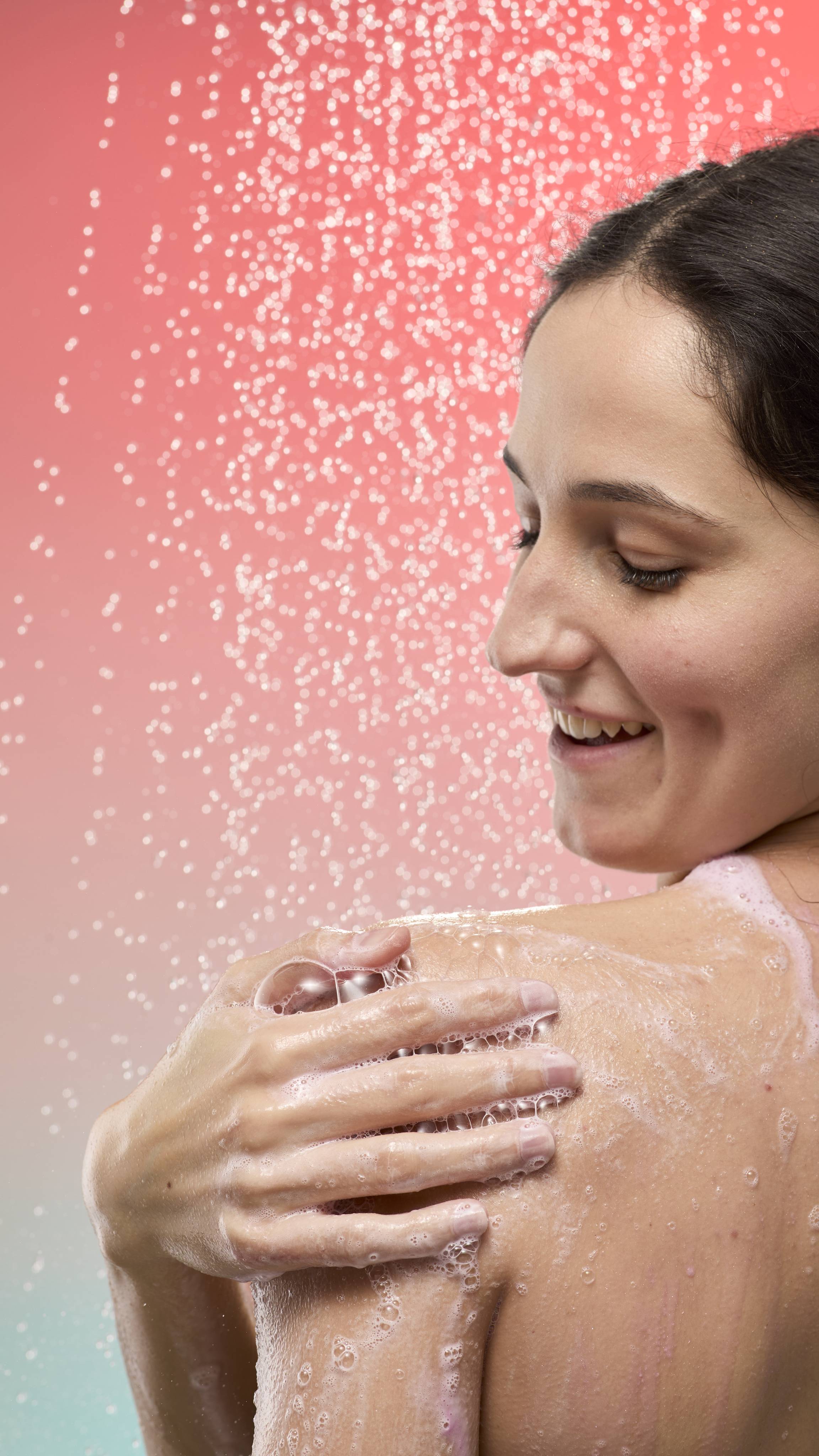 A smiling person lathers her shoulder with pale pink foam under a shower spray. Set against a peach-to-blue gradient background with fine water droplets falling.