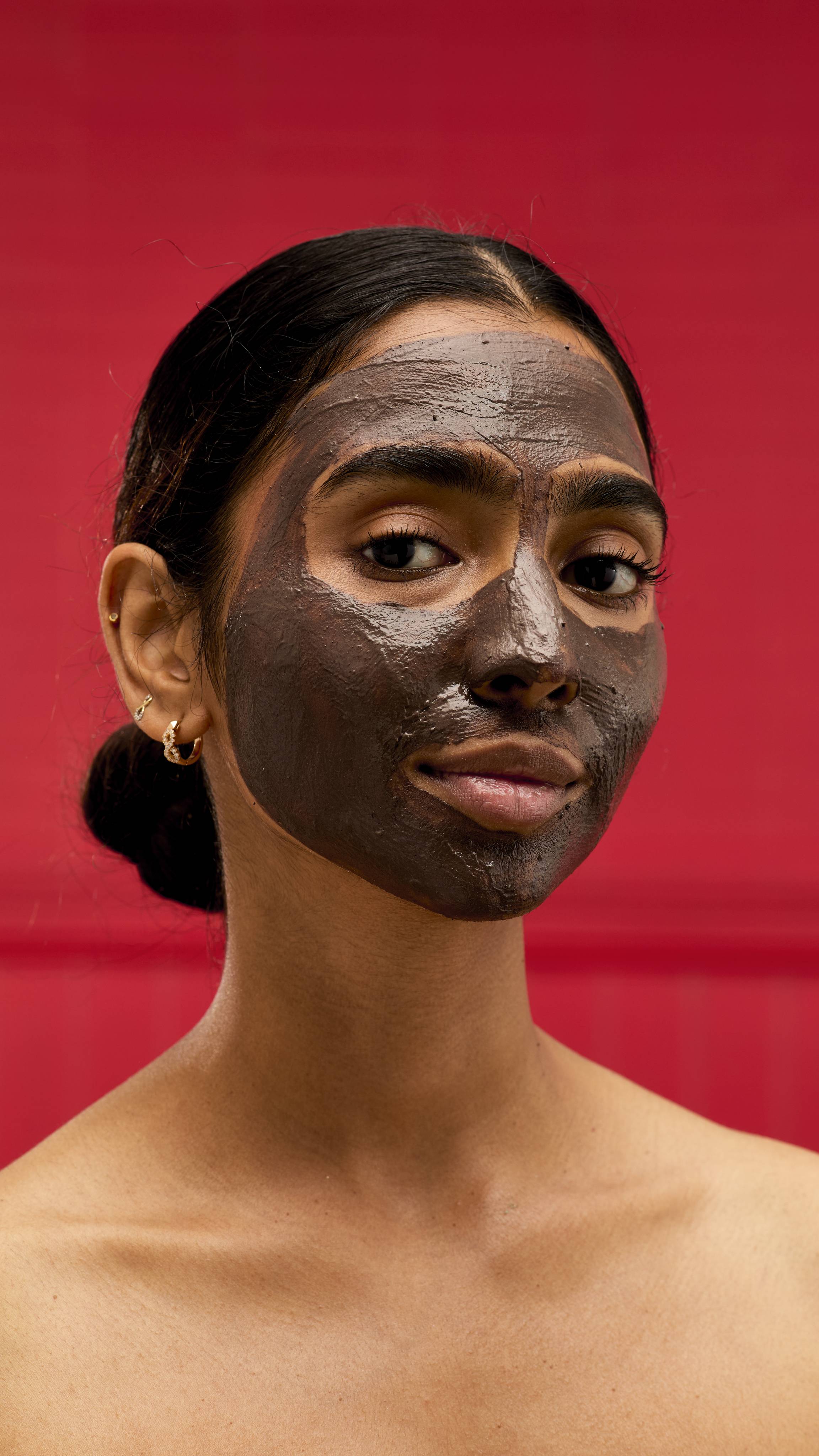 A person with their hair in a bun looks directly at the camera with a LUSH Rudolph jelly mask applied to their entire face. The dark, glossy mask contrasts with the vibrant red background.



