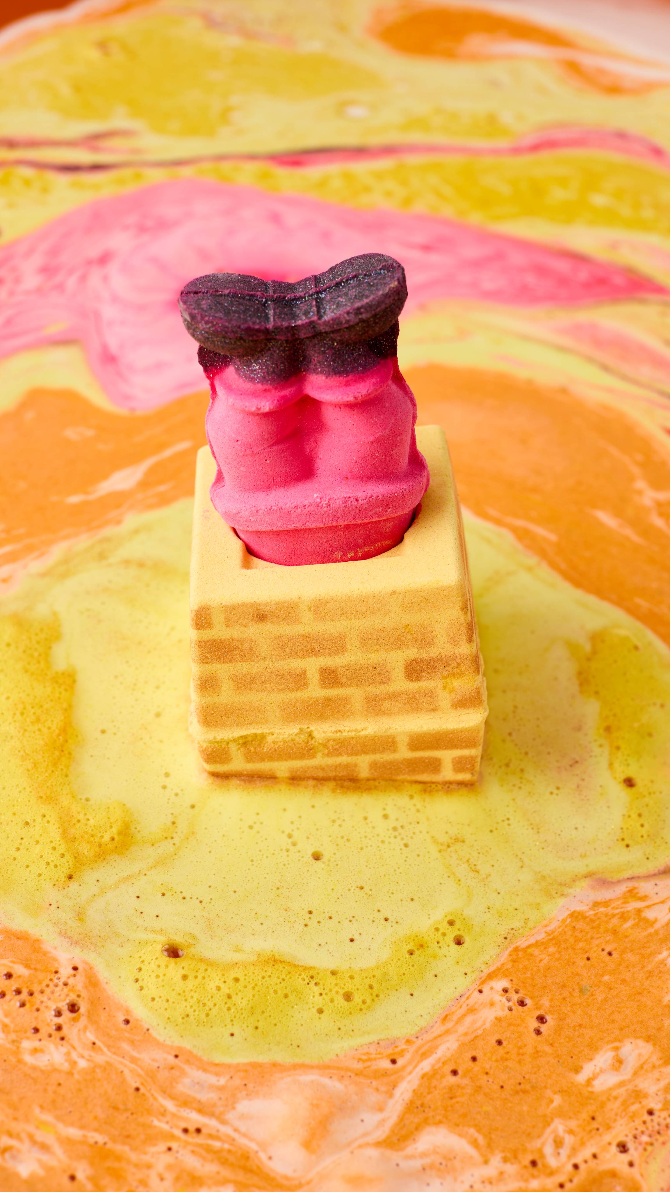 The image shows a yellow, brick-patterned Stuck In The Chimney bath bomb fizzing in swirling yellow and orange bath art. A removable figure's red legs and black boots stick out of the top as ripples of colourful foam surround it.