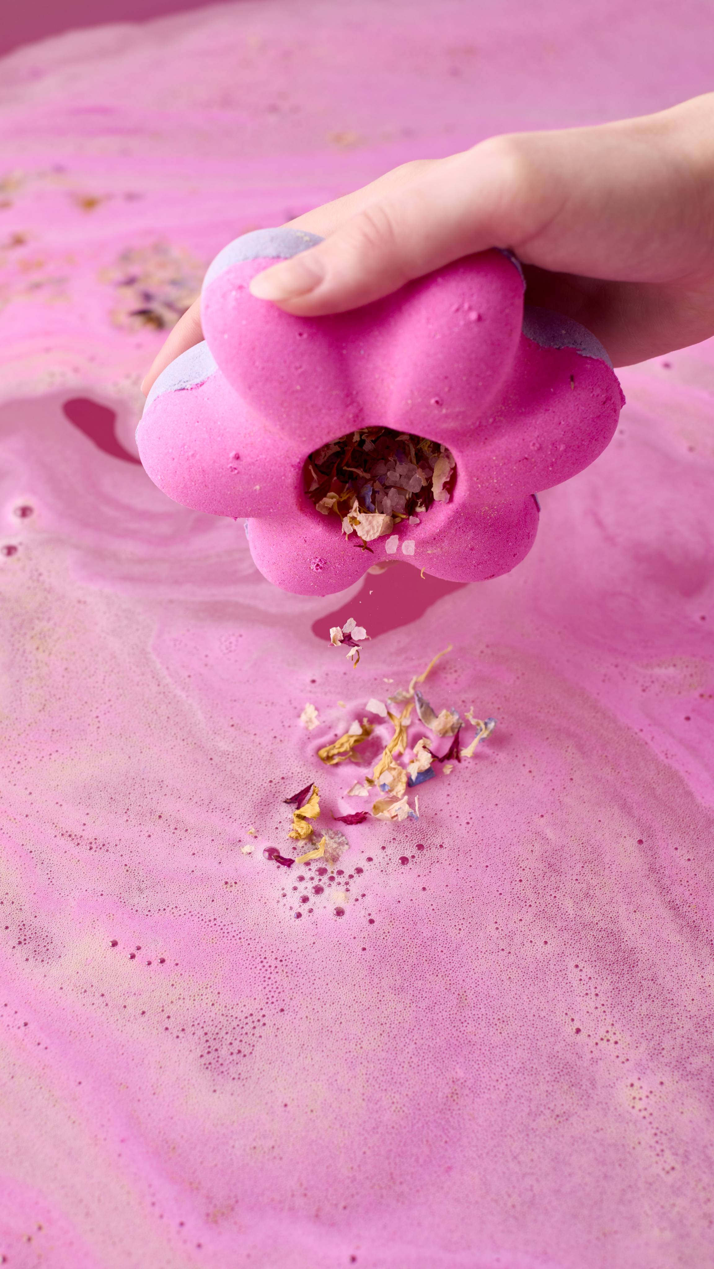 A hand holds the magenta Secret Garden Bombshell bath bomb upside down, sprinkling dried flower petals and salts from its hollow center into the vibrant pink bathwater below.