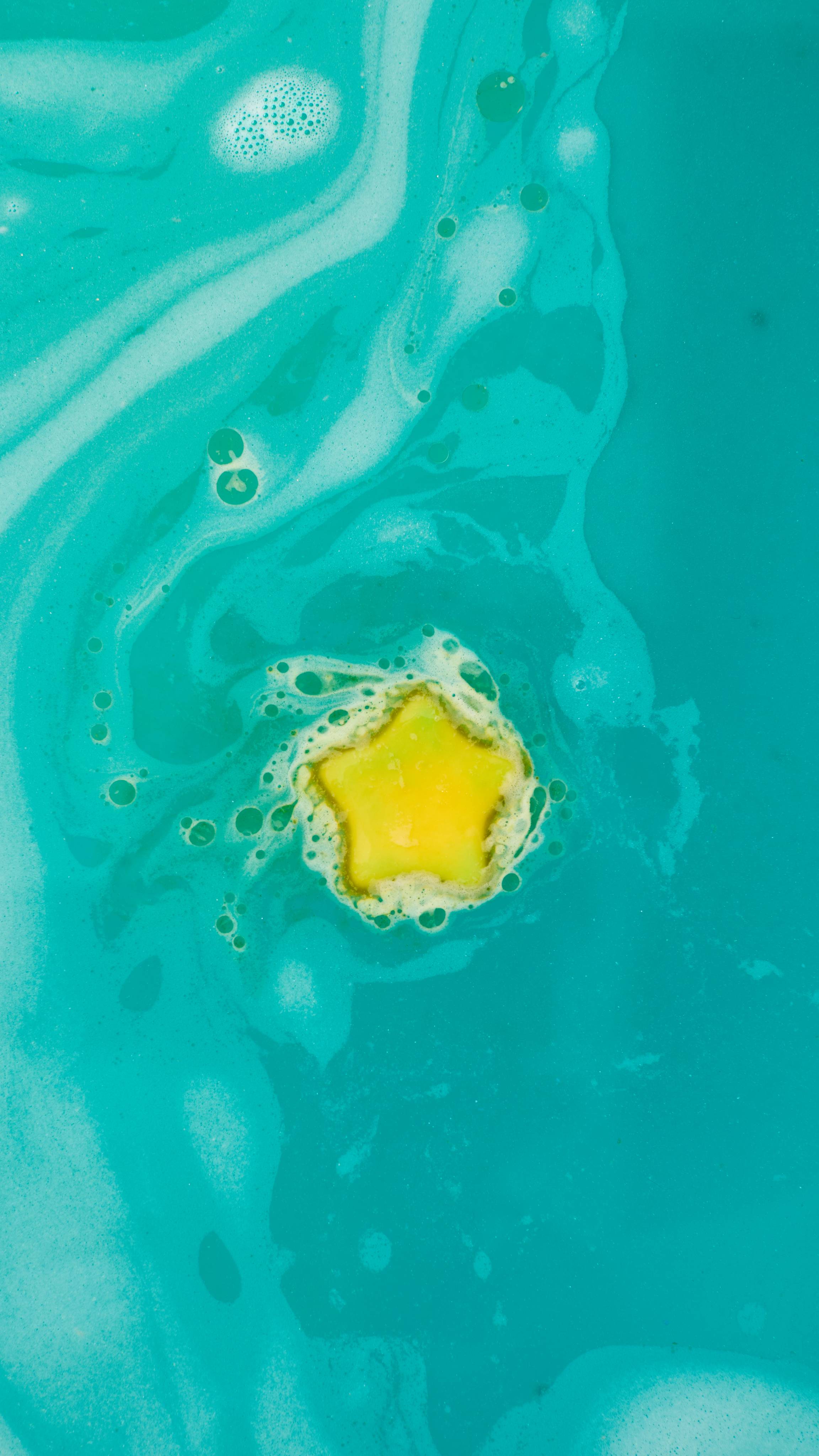 The image shows a vibrant yellow, star-shaped bath melt topper of the 'Shoot For The Stars' bath bomb floating in the water. Hues of dark and light turquoise swirl around the product, mixing with abundant foam and bubbles.