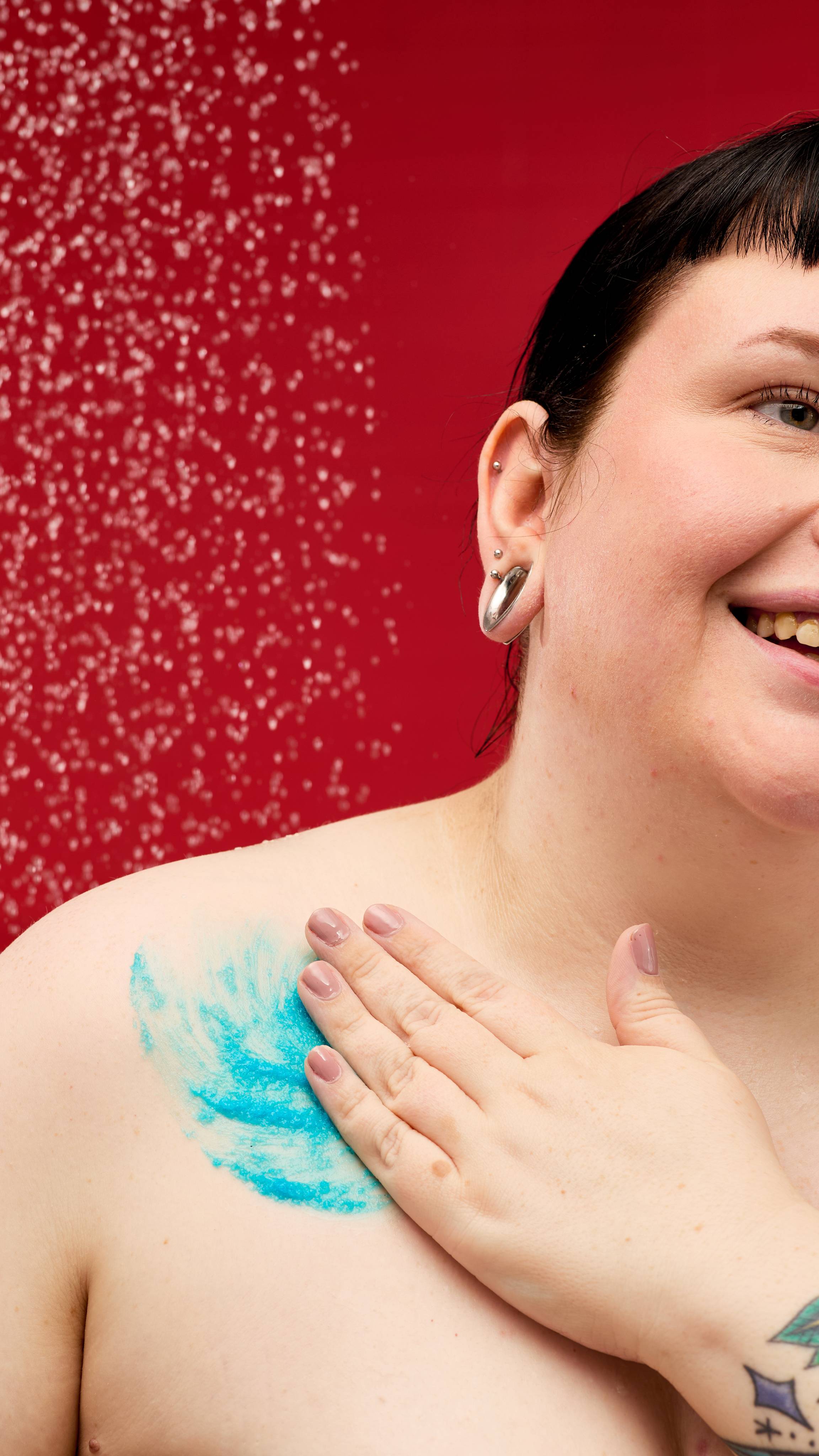 A person smiles as they apply a bright blue, chunky LUSH Shoot for the Stars body scrub to their collarbone and shoulder. Water droplets are visible in the air, creating a starry effect against a vibrant red background.
