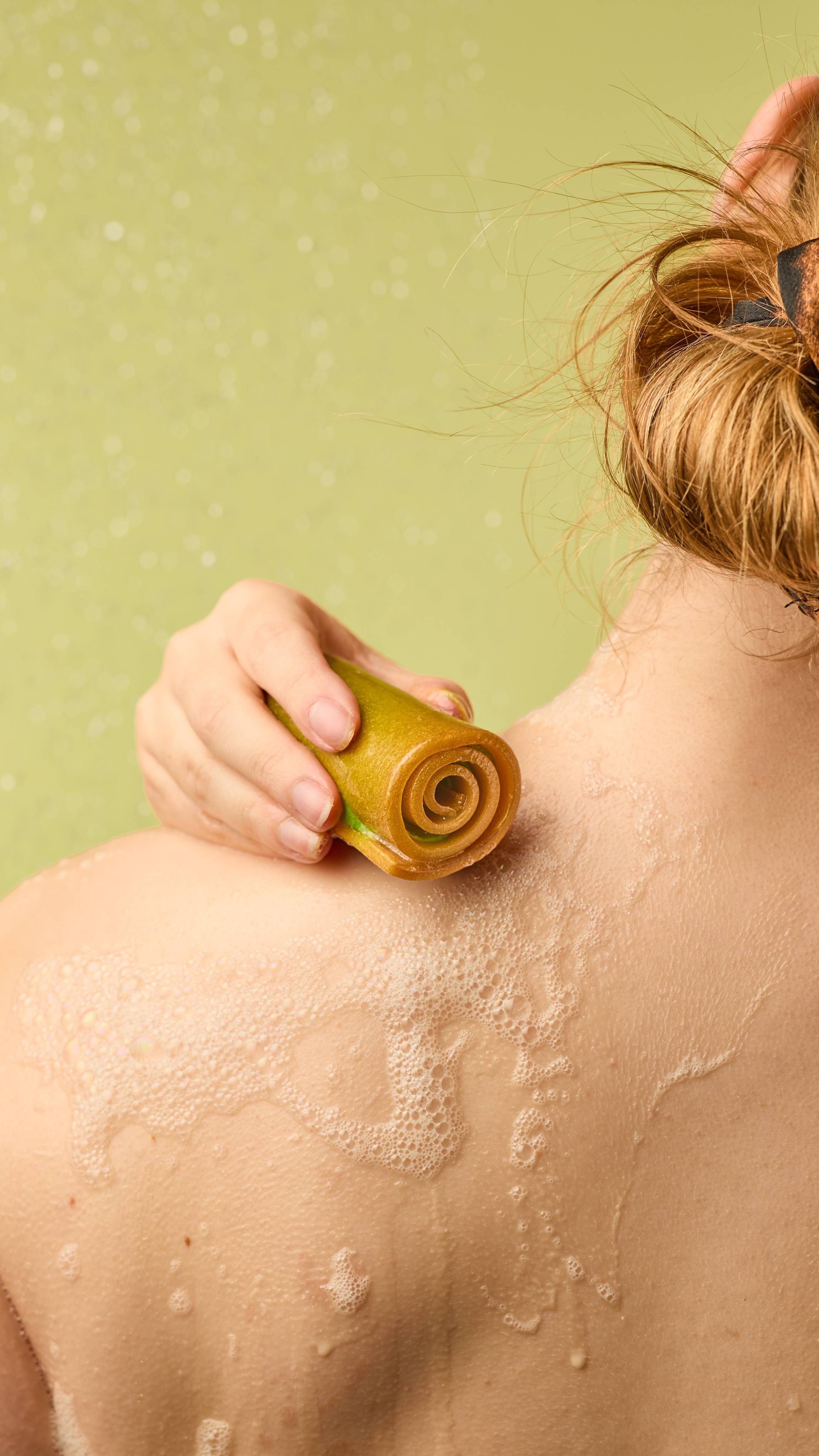 The image shows a person rubbing a cylindrical, caramel-coloured Showstopper soap with golden lustre over their neck and shoulder. White, bubbly lather forms on the skin, against a light green background.