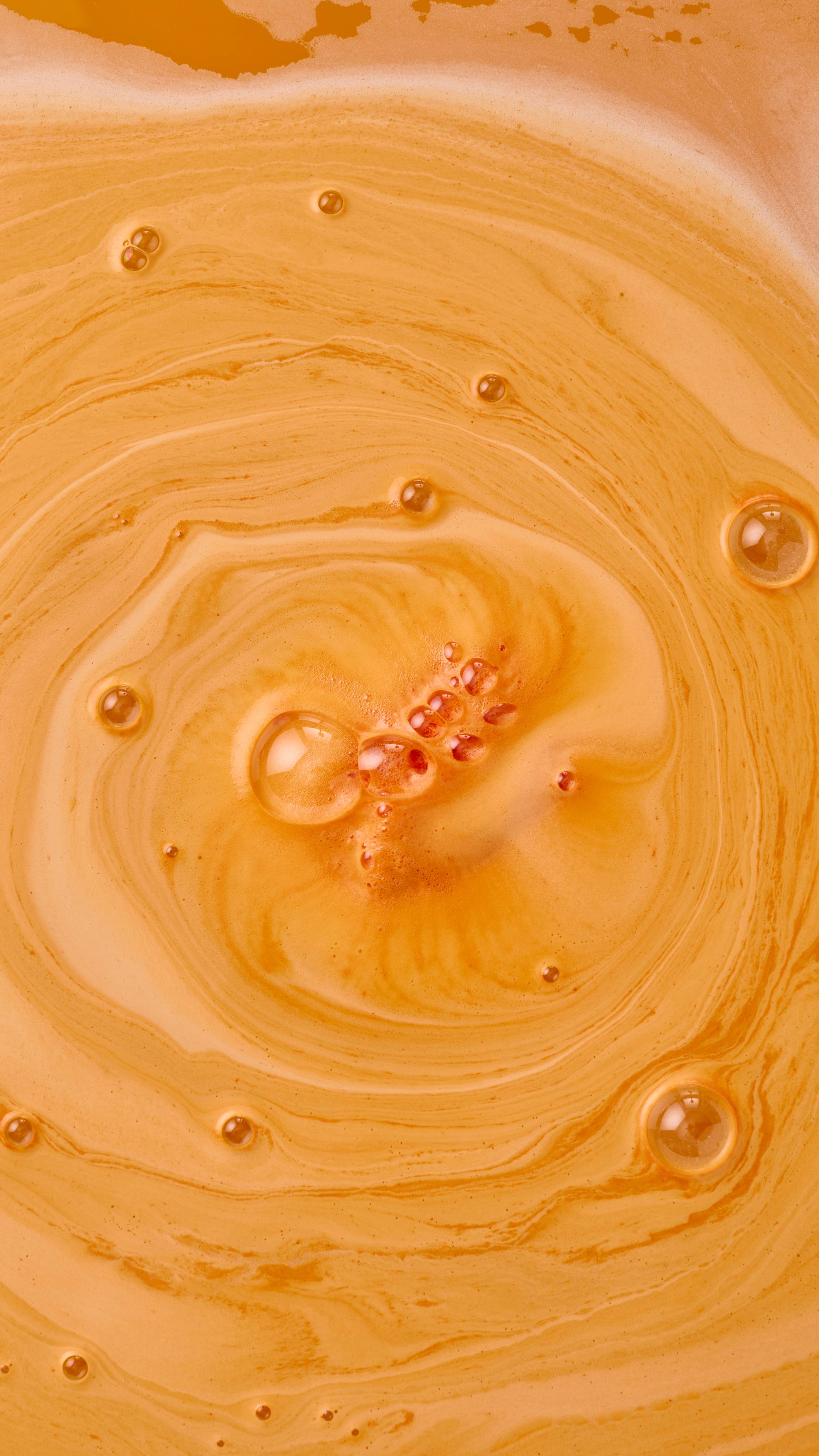 An aerial view of a bathtub filled with bright orange water and swirling, creamy foam, created by the "Shrimply The Best" bath bomb. Large, clear bubbles with iridescent reflections float on the surface of the velvety, orange foam.