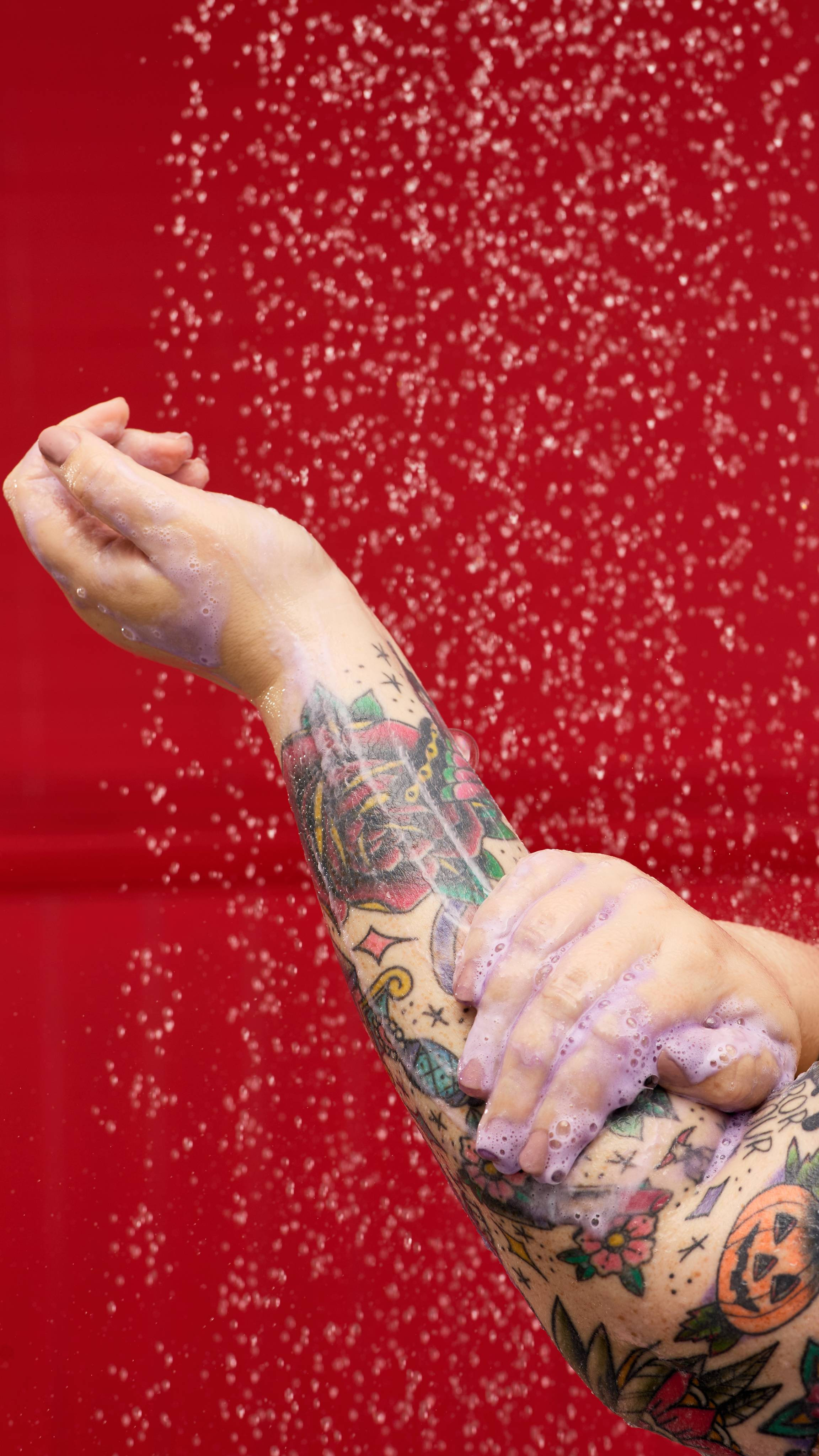 A pair of tattooed forearms is lathering up with a foamy, purple-hued product, suggesting the use of a LUSH Sleepy Bear shower gel. Water droplets are visible in the air against a vibrant red background.
