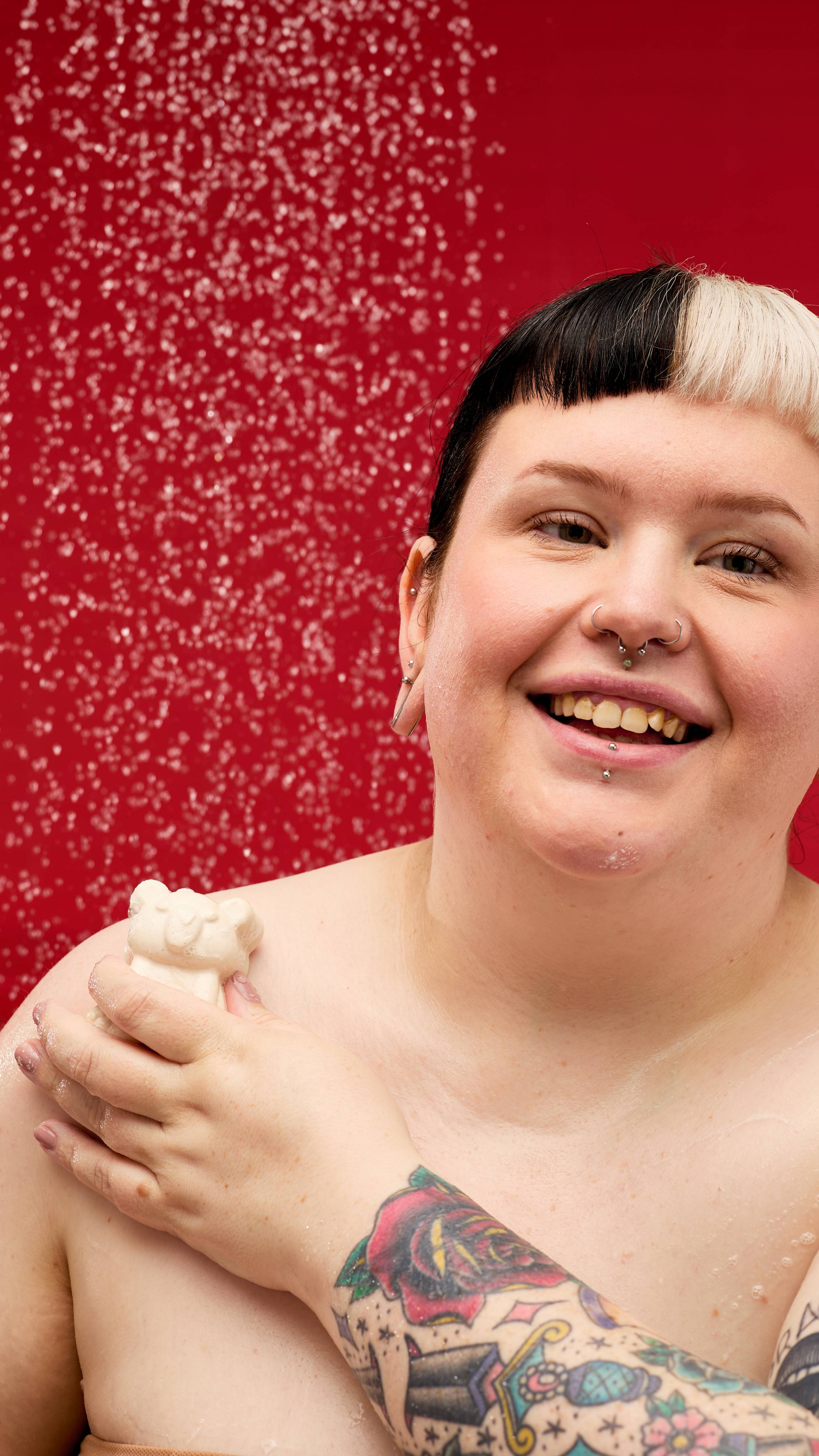 A person is shown from the chest up, in a shower against a red background. They're smiling and looking up, with water droplets on their face and body. They are using the white Snow Bear soap to wash their shoulder.