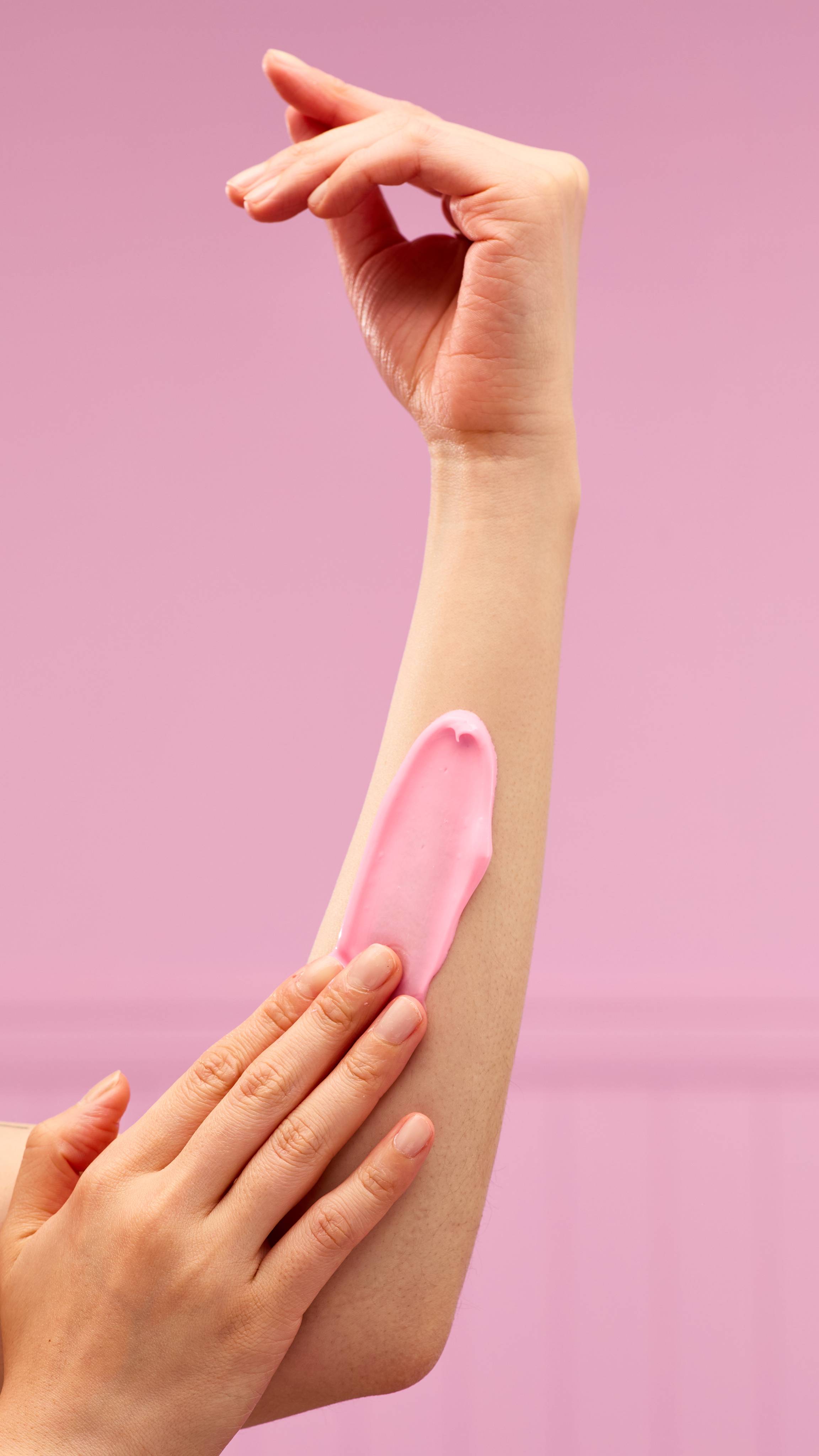 A hand is applying a thick, creamy swirl of LUSH Snow Fairy body lotion to a forearm, with the textured product standing out against a vibrant pink background.