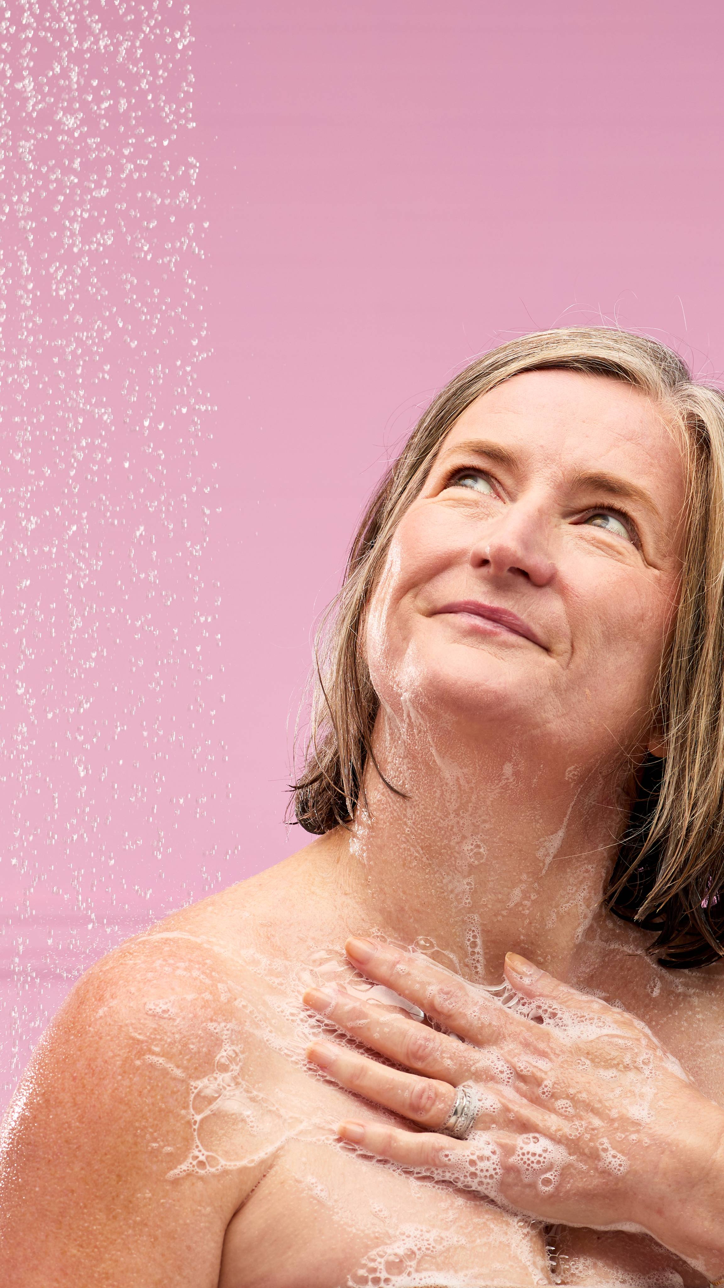 A person is shown from the chest up, in a shower against a pink background with blurred, shower water. They're smiling and looking up, as they use the Snow Gairy Glitter shower gel to create a rich lather on their shoulder. 