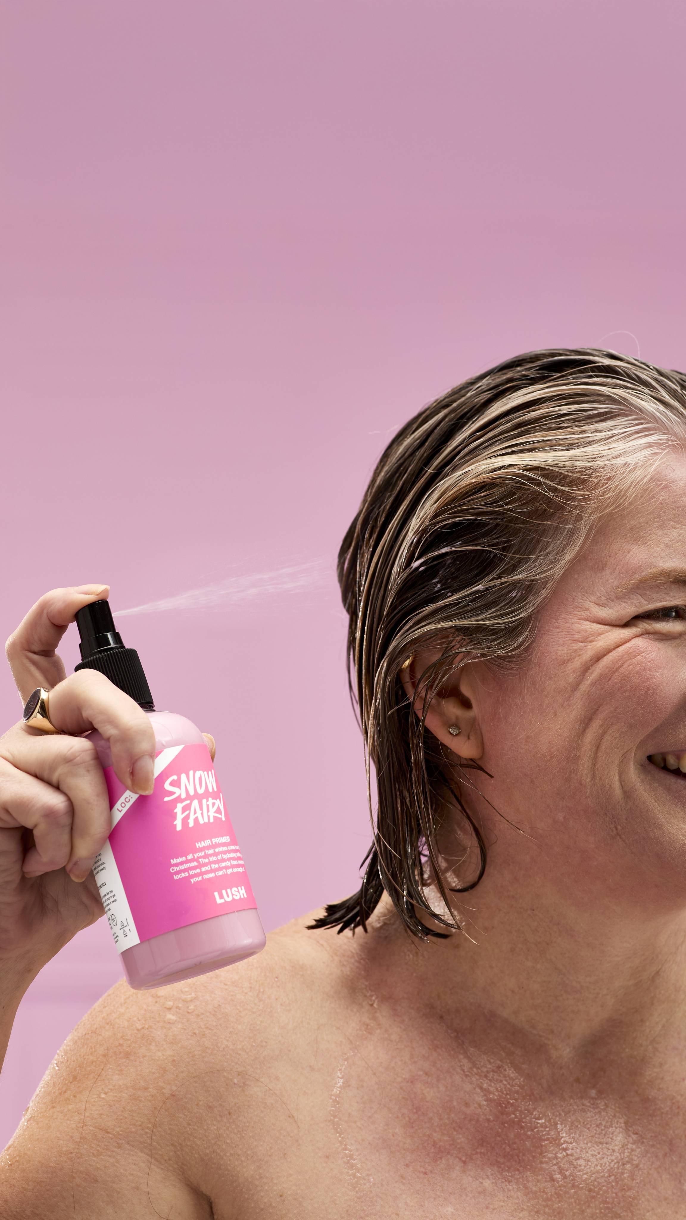A person smiles while spritzing LUSH Snow Fairy hair primer onto their wet hair. The pink bottle, with its product information visible, is held in one hand. The background is a soft pink.