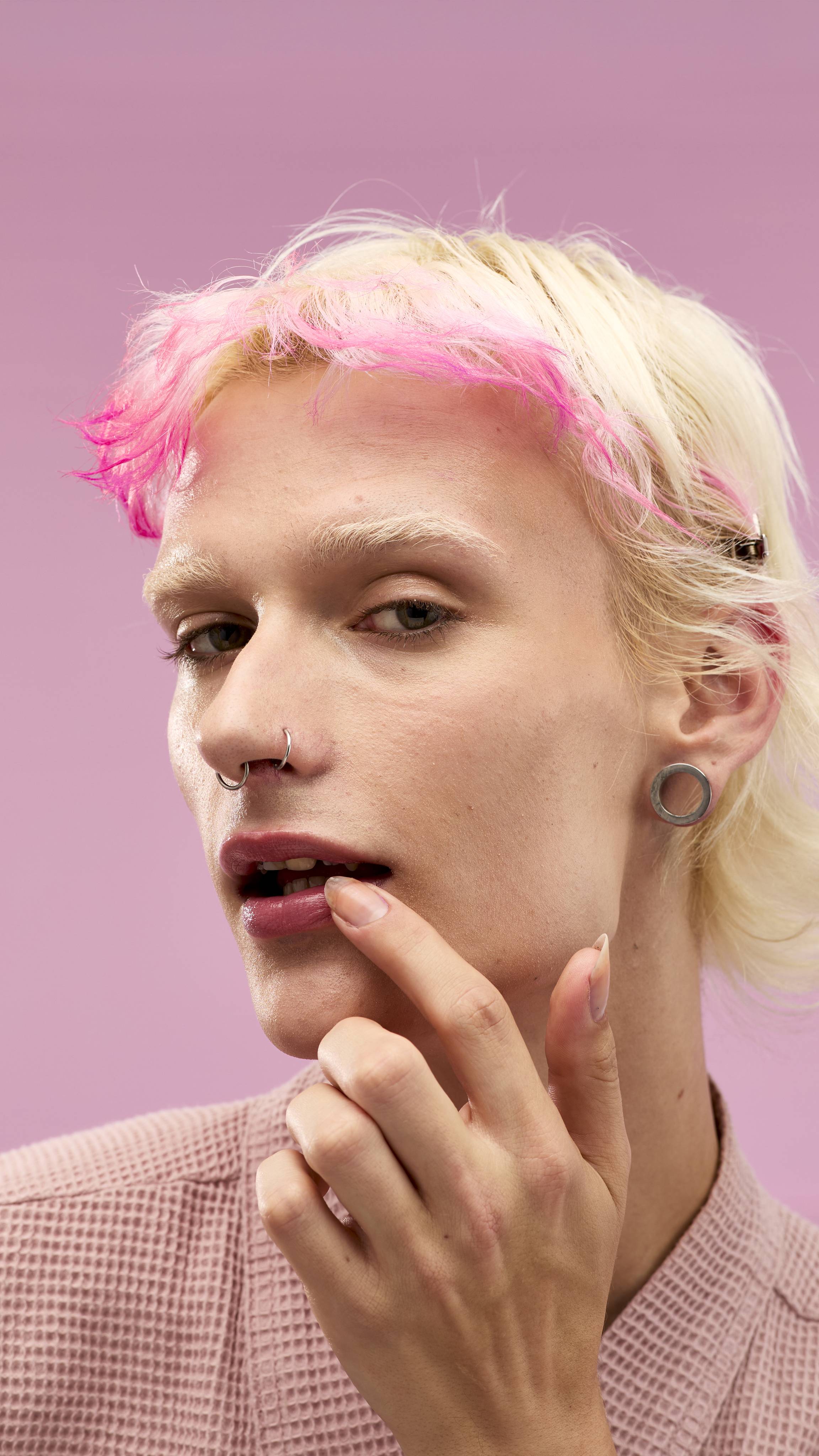 
A person with blonde hair and vibrant pink bangs looks at the camera while touching their glossy lips with a finger. The lips have a subtle pink sheen, suggesting the use of a LUSH Snow Fairy lip mask. The background is a soft pink.