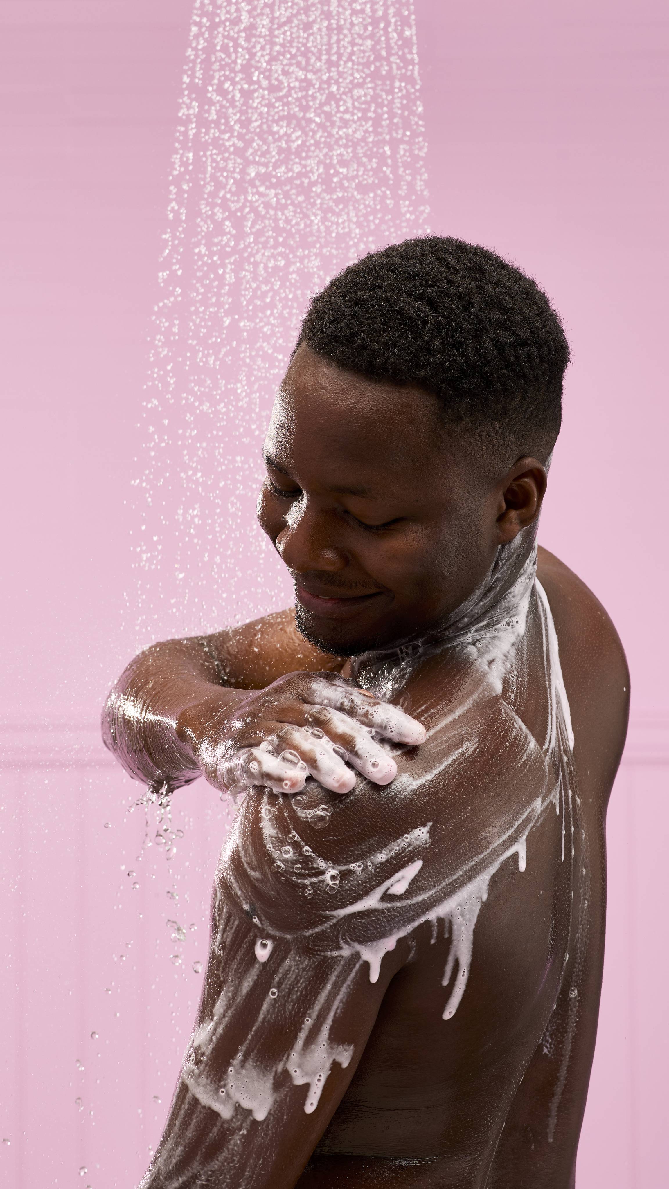 A person with dark skin is shown from the chest up, in a shower against a pink background. They're smiling and looking away from the camera as they rub the Snow Fairy Milkshake shower gel into white lather over their shoulder.