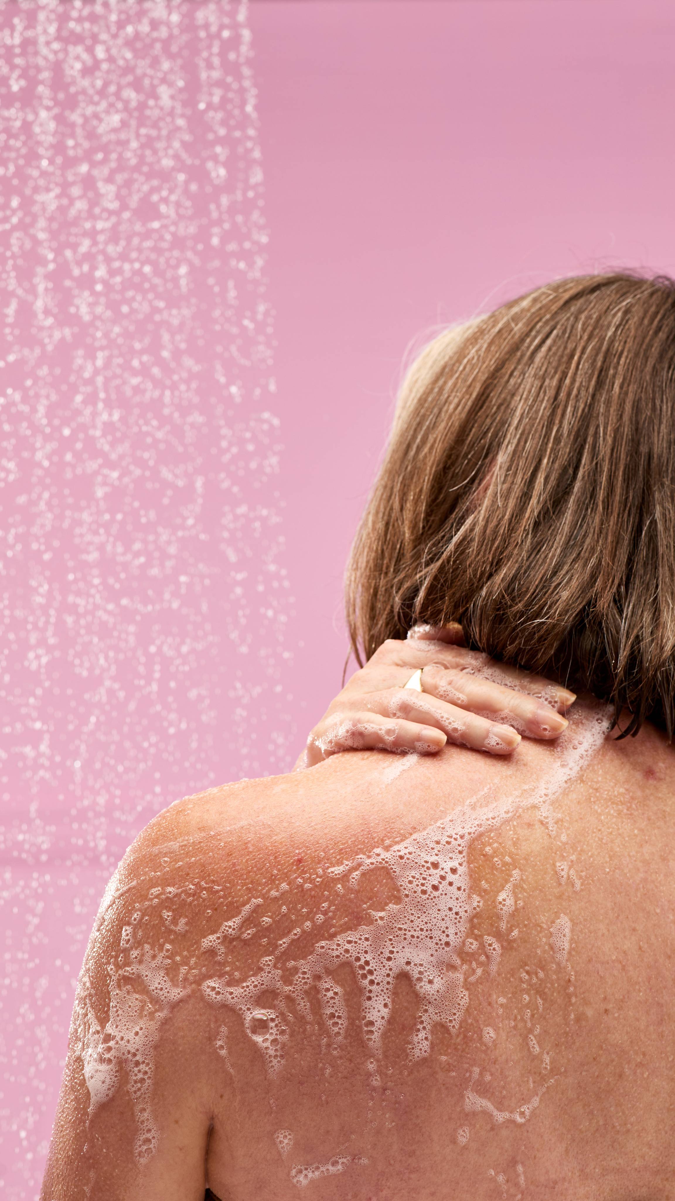 A person's hand with a ring on it is shown from behind, lathering a foamy, vibrant pink LUSH Snow Fairy shower gel on their shoulder. Bubbles are visible on the skin and water droplets sparkle in the air against a soft pink background.