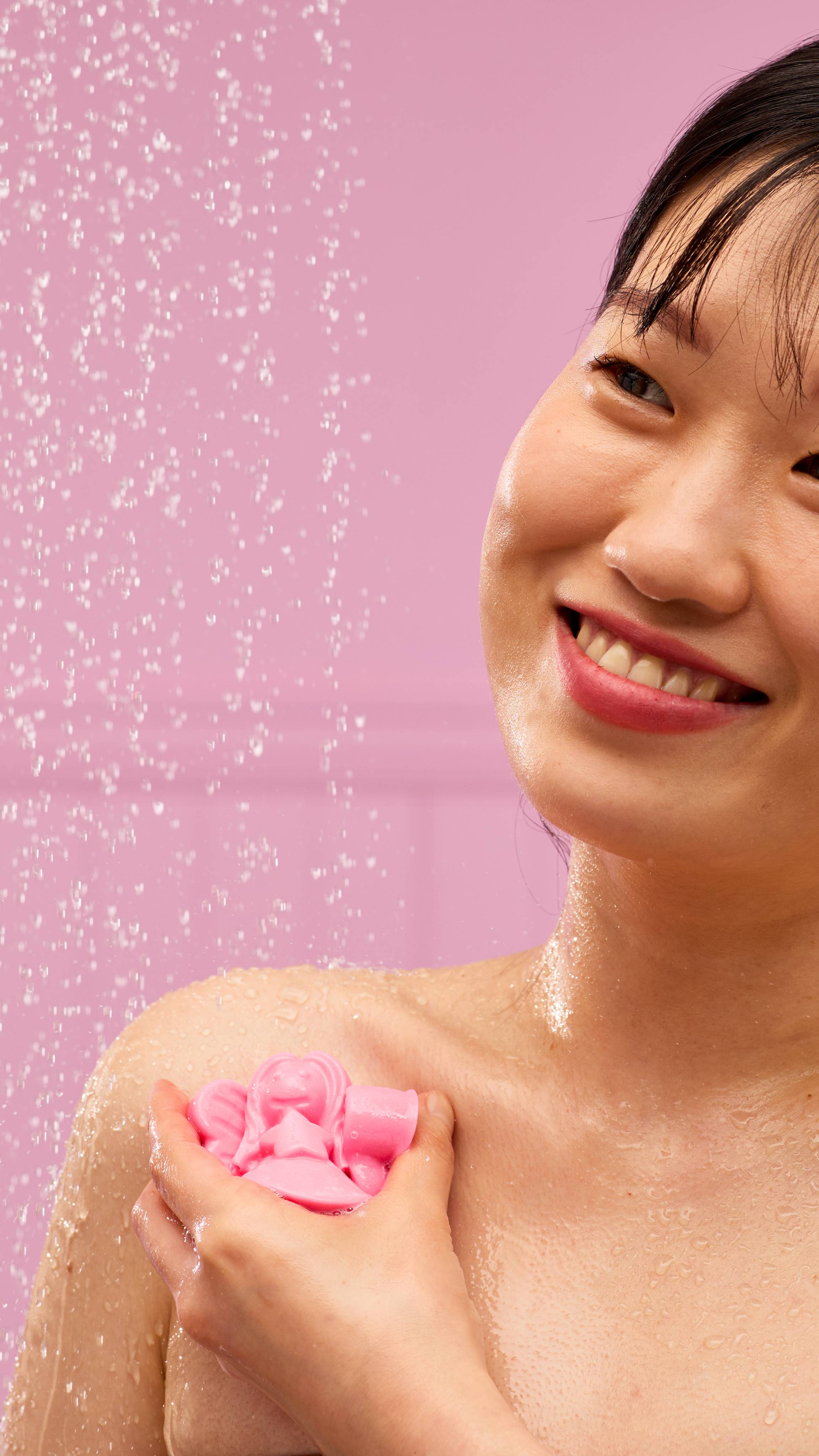 A person smiles while holding a solid, pink, angel-shaped LUSH Snow Fairy shower jelly on their shoulder. Water droplets glisten on their skin and are visible in the air against a soft pink background.
