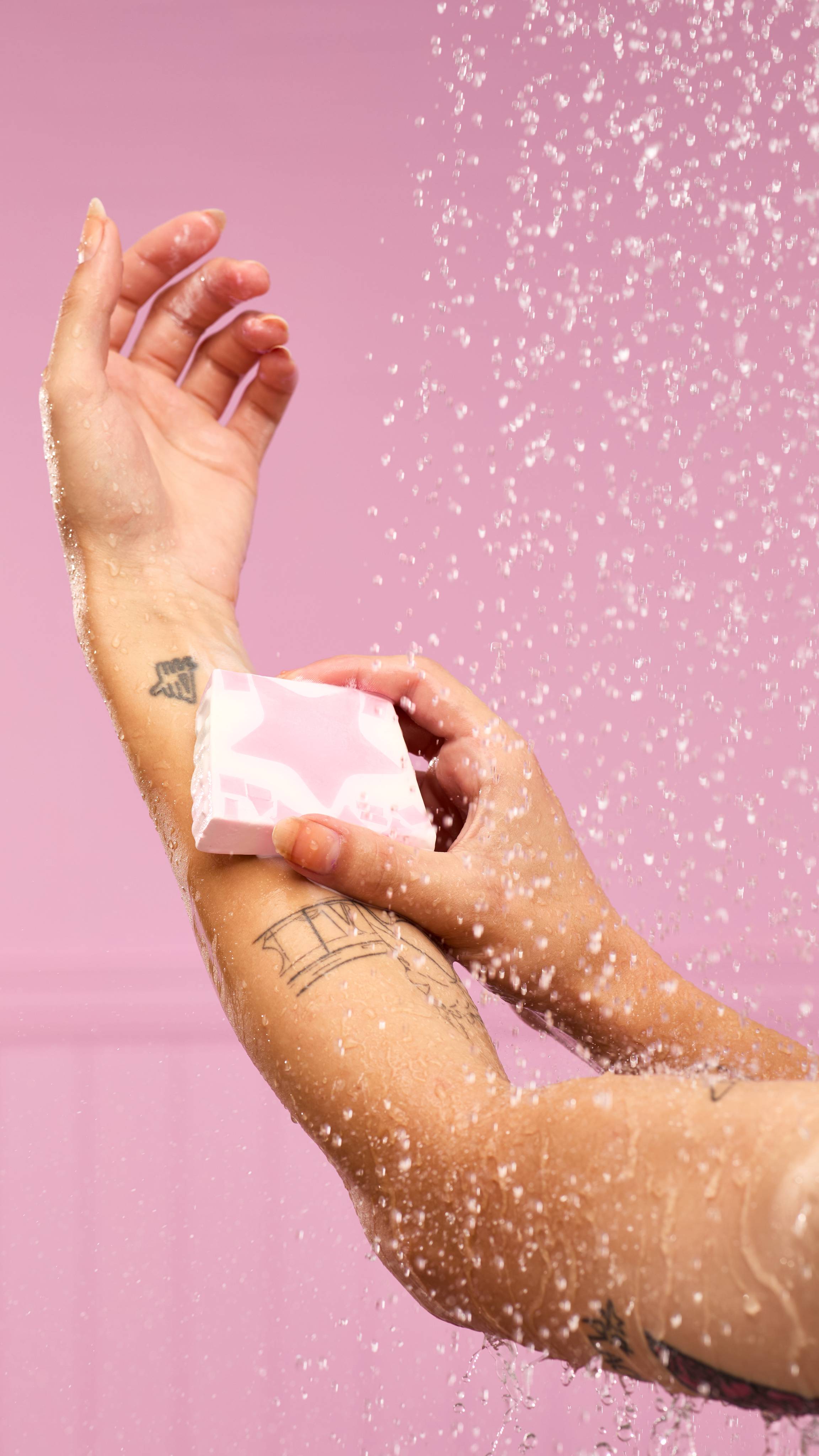 A fair-skinned arm with a wrist tattoo is in a shower. Water streams down, and they are holding the pink Snow Fairy soap slice with a pink star at its centre. They are rubbing the soap on their forearm.