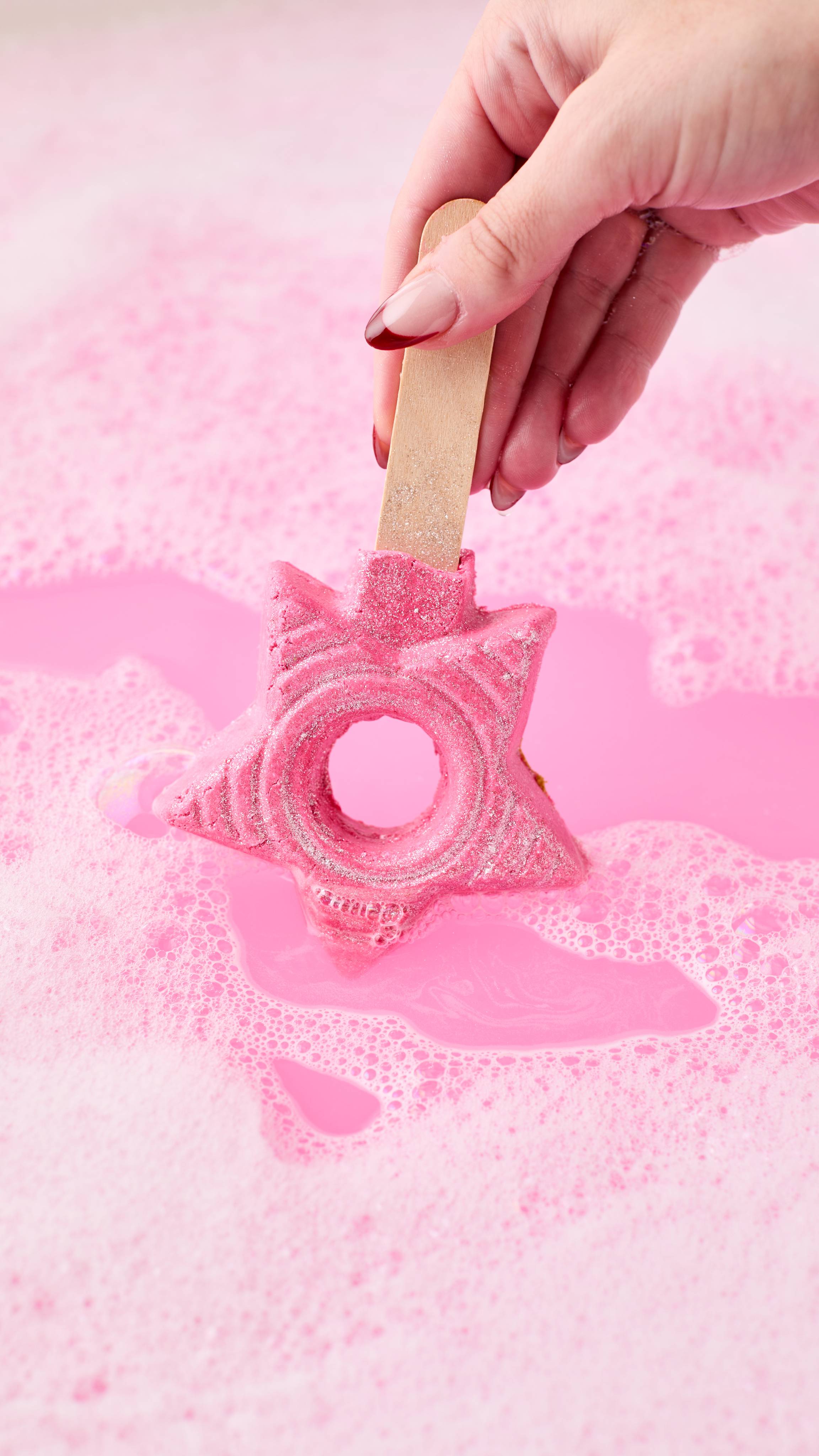 The image shows a hand stirring a vibrant pink, star-shaped Snow Fairy Bubble Wand through bright pink bath water. The glittery product creates a bubbly, swirling foam around the wand and stick.