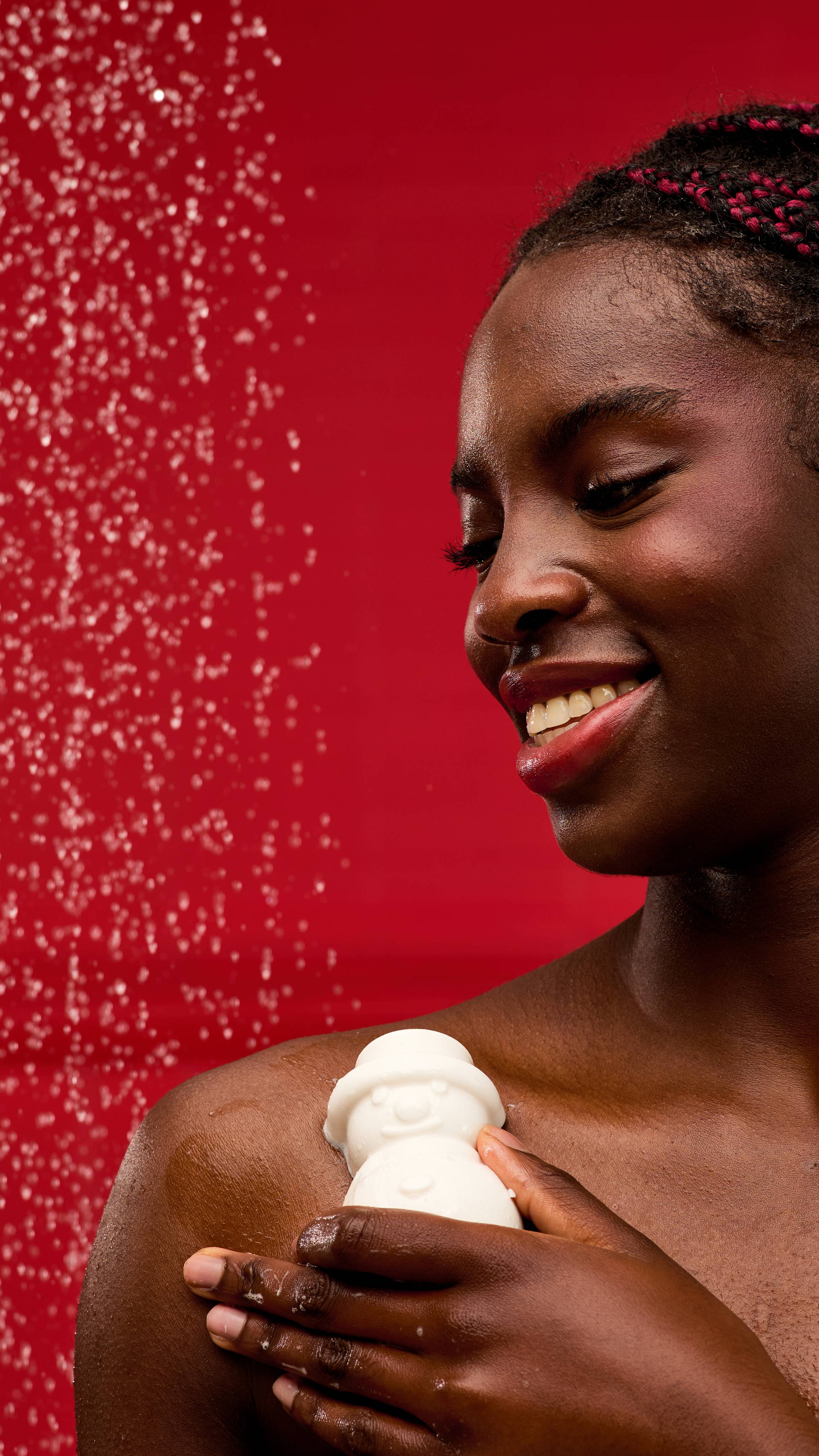 A person smiles as they use a solid, white, snowman-shaped LUSH Snowy shower gummy on their shoulder. The skin is wet and shiny, with water droplets visible in the air against a vibrant red background.