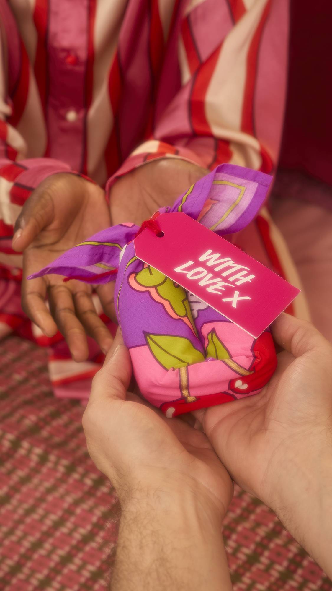 Hands pass a gift wrapped in the purple Squeeze Knot Wrap with green leaves to a person in pink striped pajamas. A pink tag reading "WITH LOVE X" faces forward.
