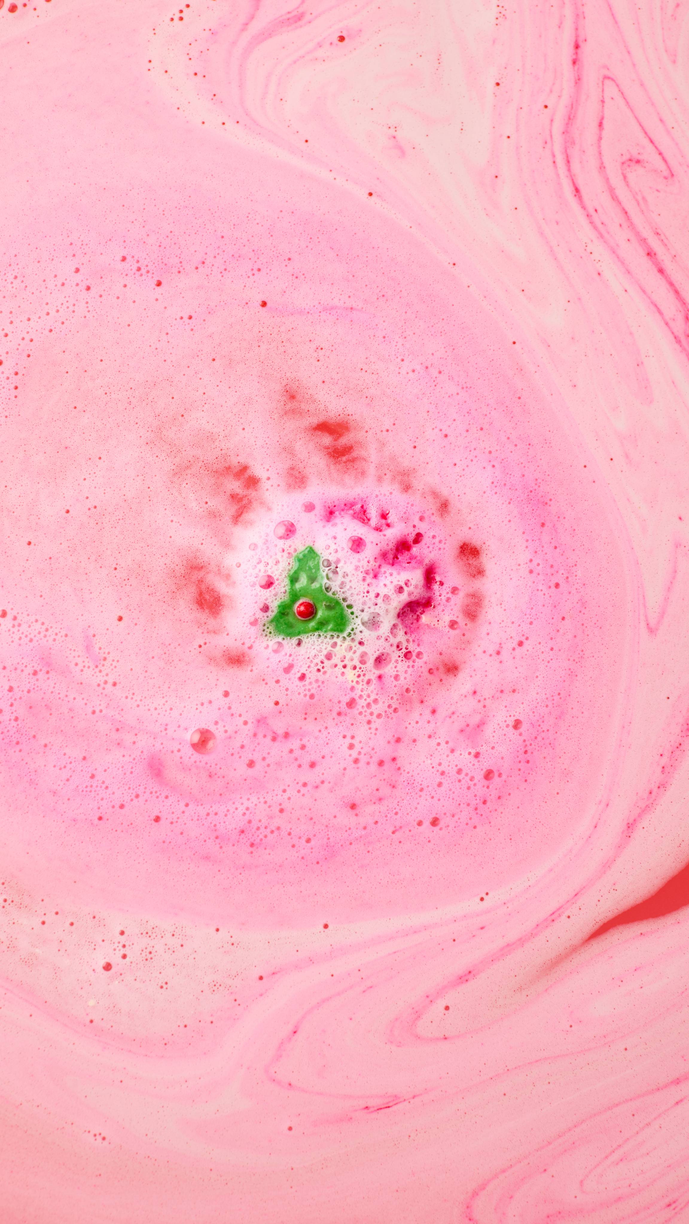 The image shows swirling bath art from the Sweet Pudding bath bomb. A small green holly leaf and red berry core remains, radiating light pink and white foam and bubbles. Darker pink swirls blend across the water.