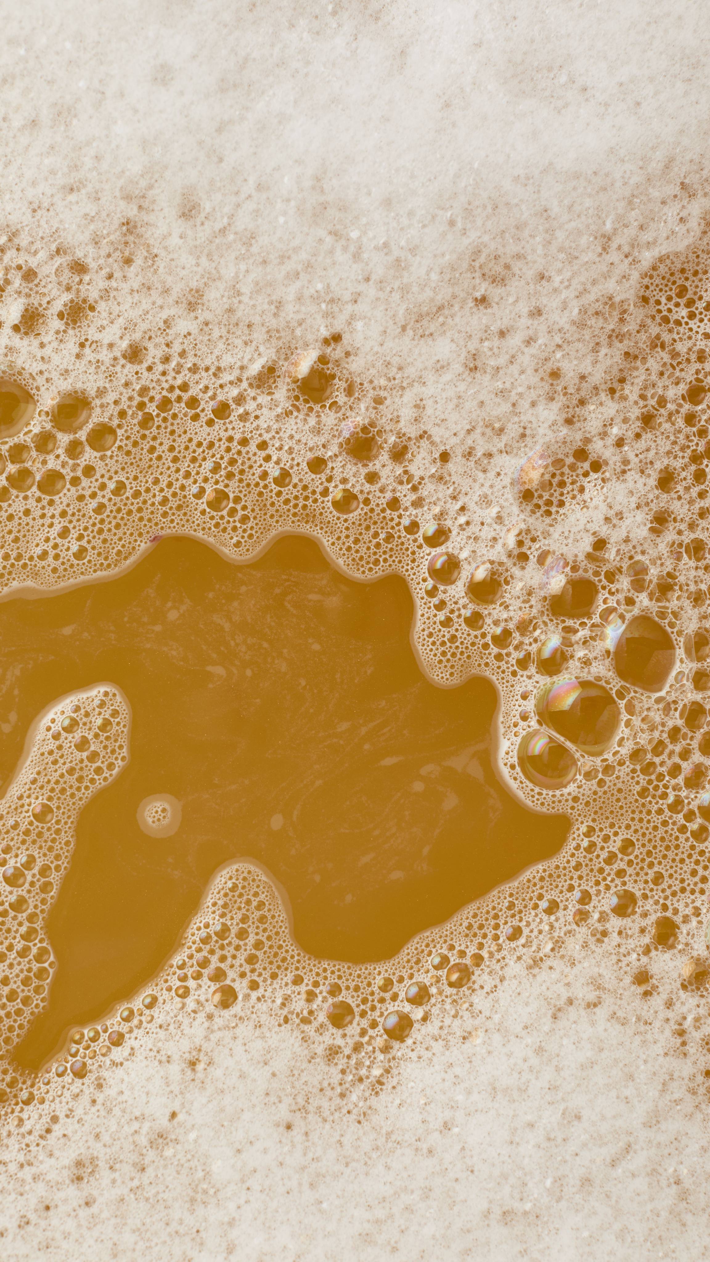A top-down view of thick, white bath bubbles over deep amber-colored water.