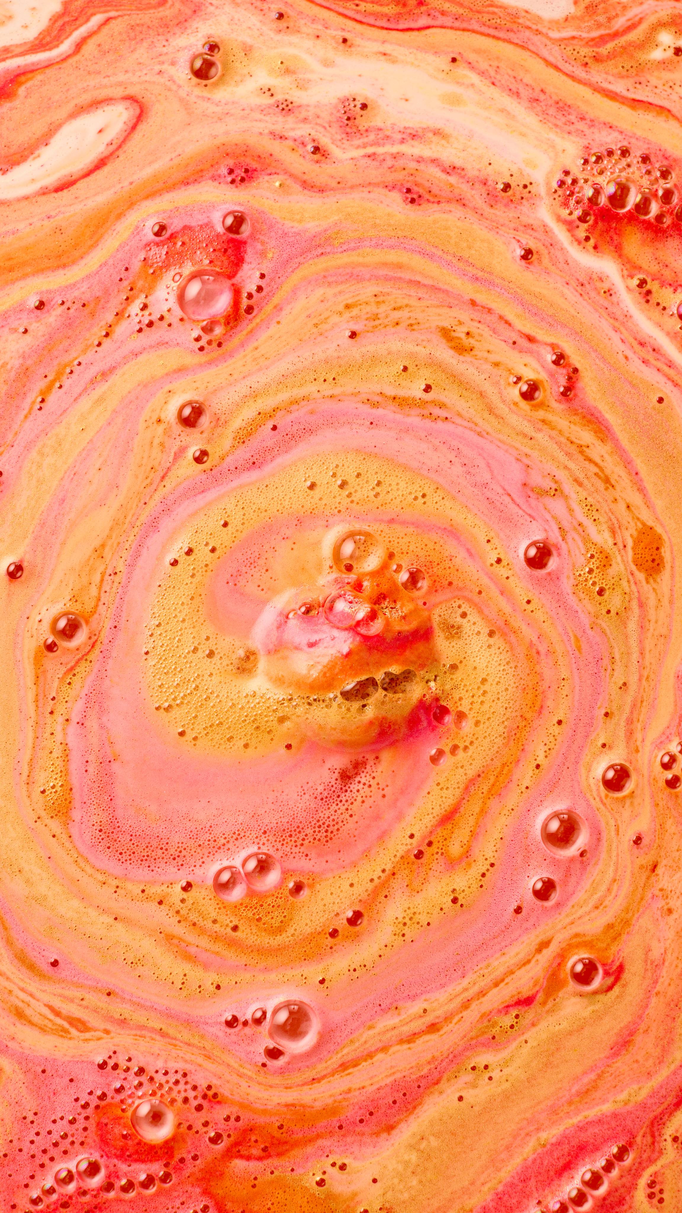 The image shows swirling bath art from 'The Alchemist' bath bomb, a rich blend of vibrant orange, yellow, and red-pink hues. Intense, creamy swirls mix with numerous prominent dark red bubbles across the water surface.