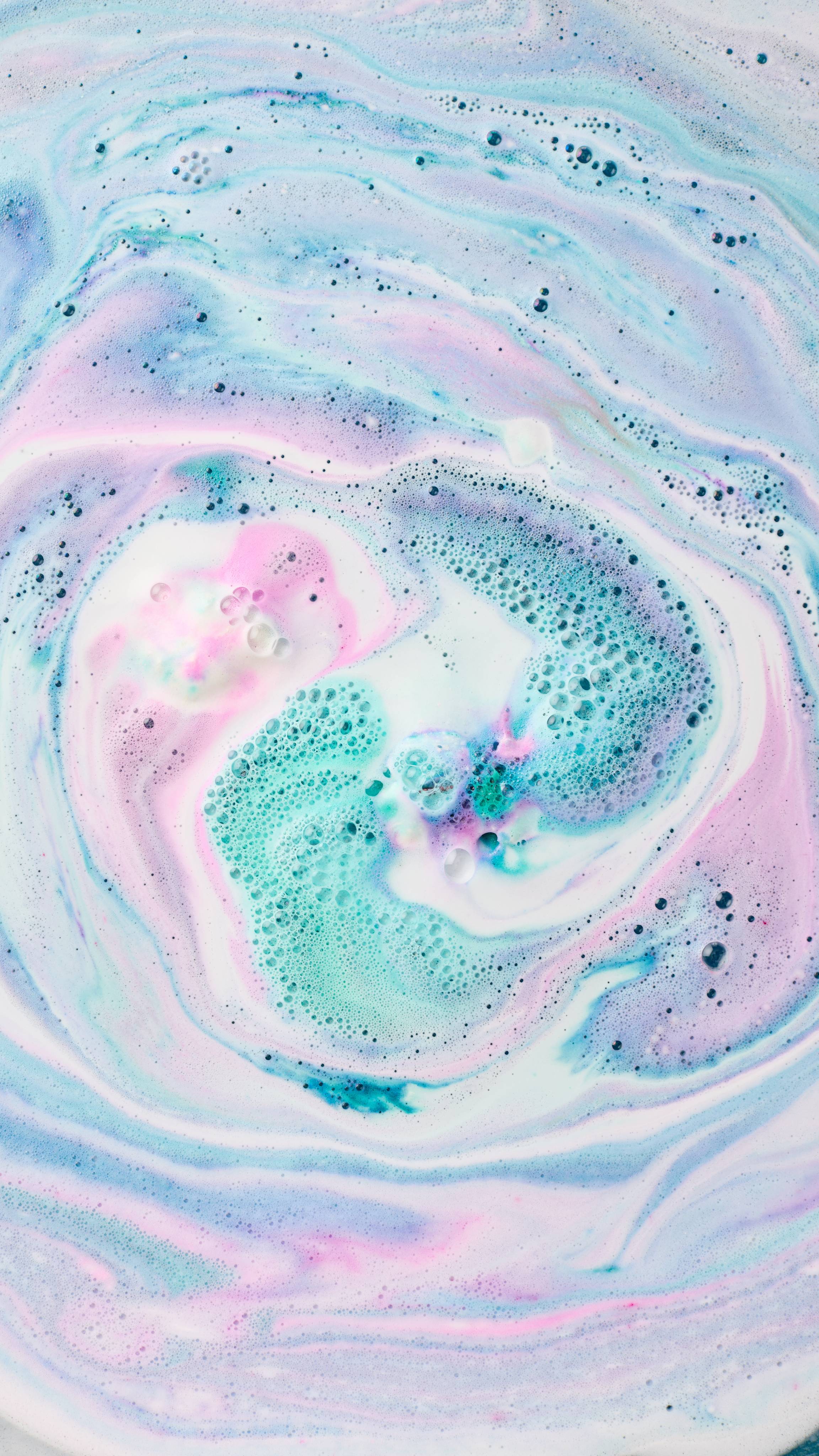 The image shows swirling bath art from 'The Girl In The Whirlwind' bath bomb, a vibrant blend of turquoise, pink, and white hues. Intricate marbled patterns mix with foam and bubbles.