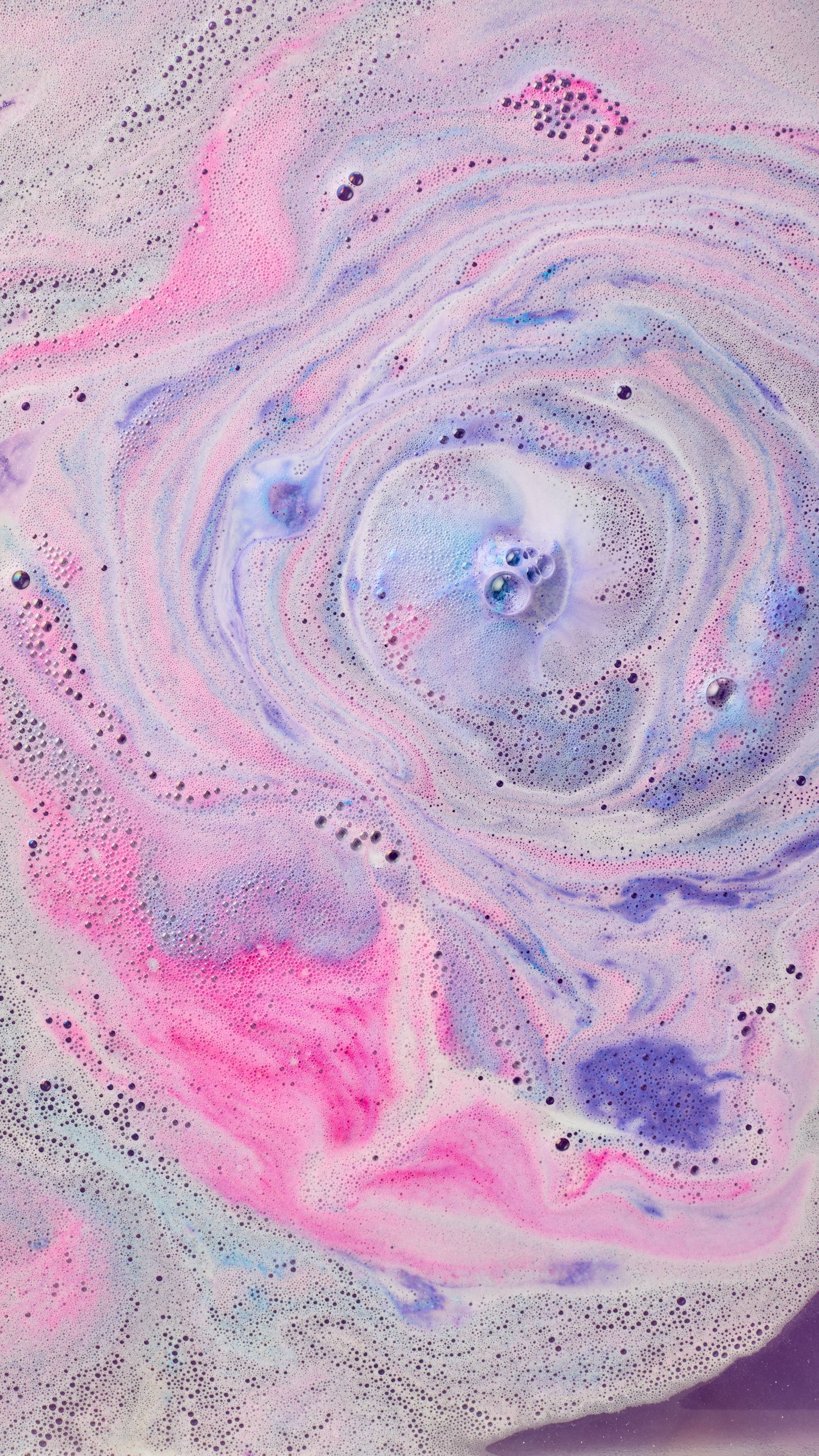 The image shows swirling bath art from 'The Magician' bath bomb, a magical blend of hot pink, purple, and light blue hues. Intense, foamy swirls radiate from the centre, mixing with scattered dark bubbles across the water.