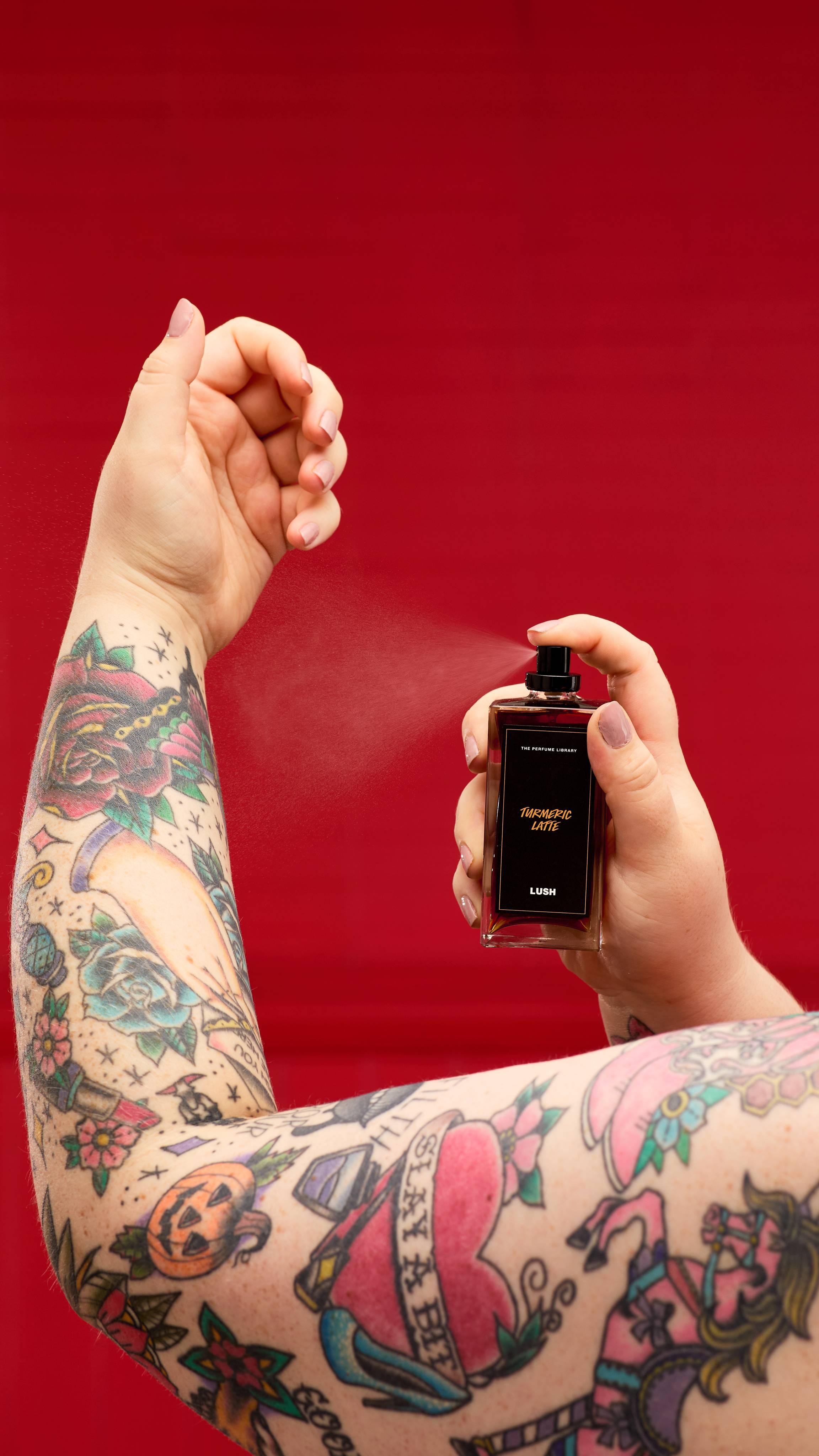 The image shows a solid, deep red background. A light-toned, tattooed arm reaches up as a hand holds the Turmeric Latter perfume bottle and spritzes the wrist and forearm. 