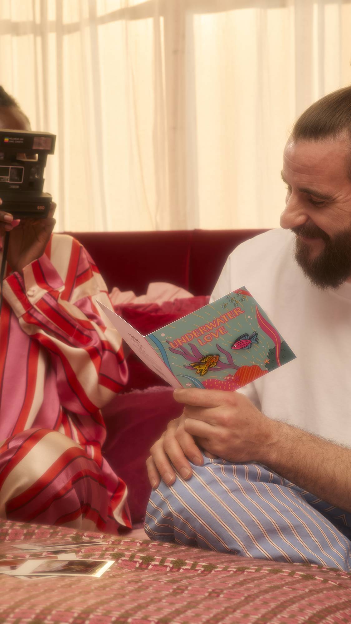 A person smiles as they read the open Underwater Love card. Beside them, a companion wearing pink and red striped pajamas holds an instant camera up to their eye to capture the moment.