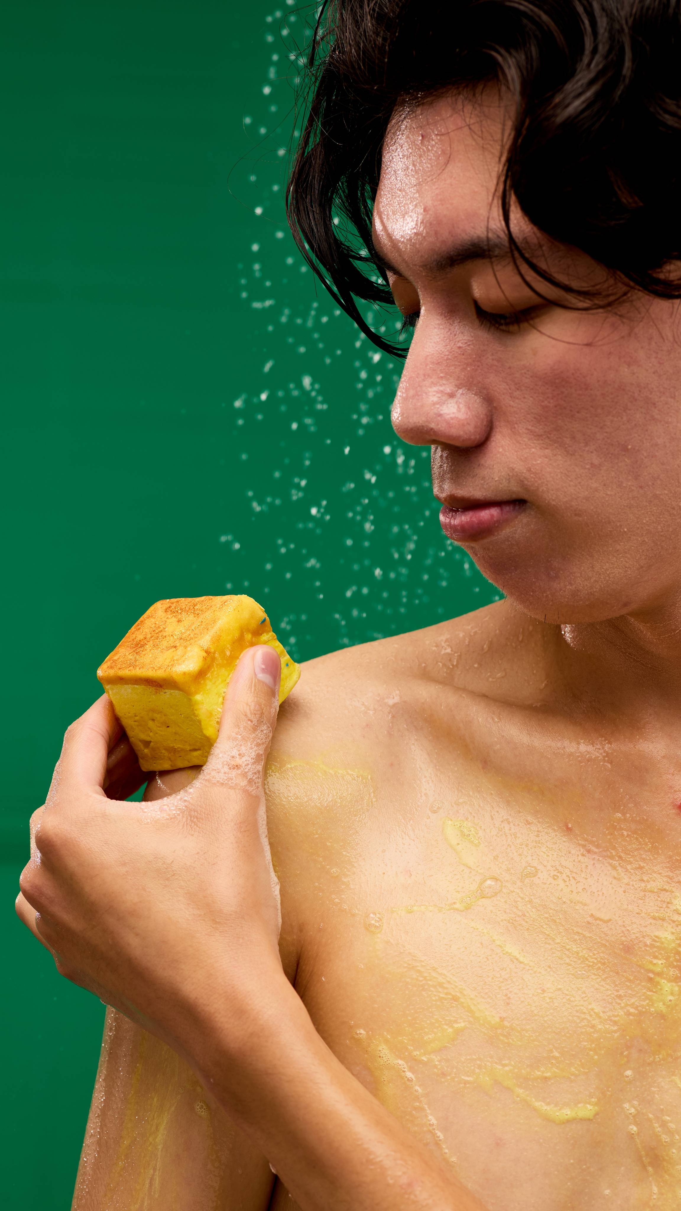 A person with wet hair lathers their chest with a square LUSH Vanilla Delight body wash bar. The pale yellow product leaves a foamy residue on their skin, with water droplets visible in the air against a vibrant green background.