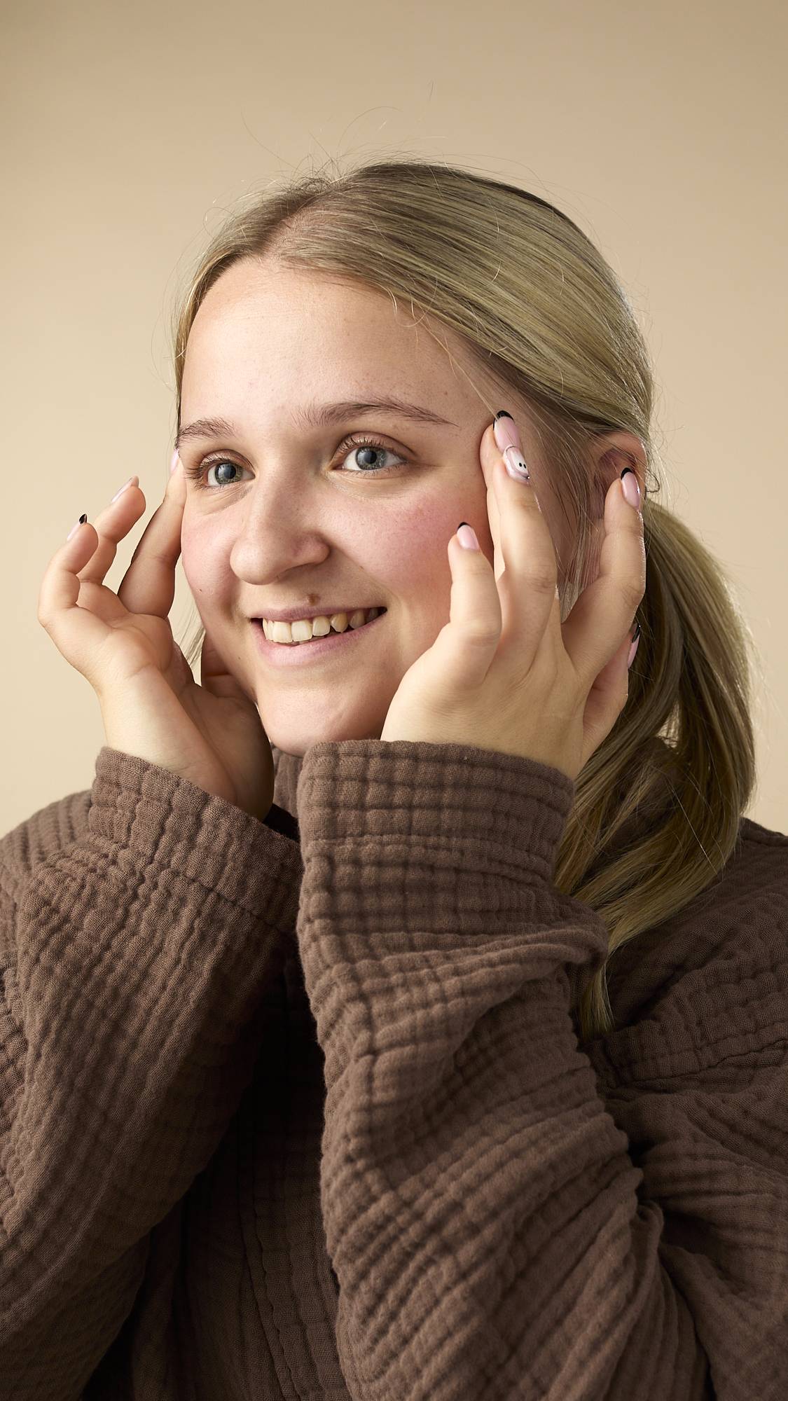 The image shows a light-skinned model with blonde hair pulled back, smiling while applying the Vision facial serum to both sides of their face. They're wearing a brown, textured robe on a light beige background.