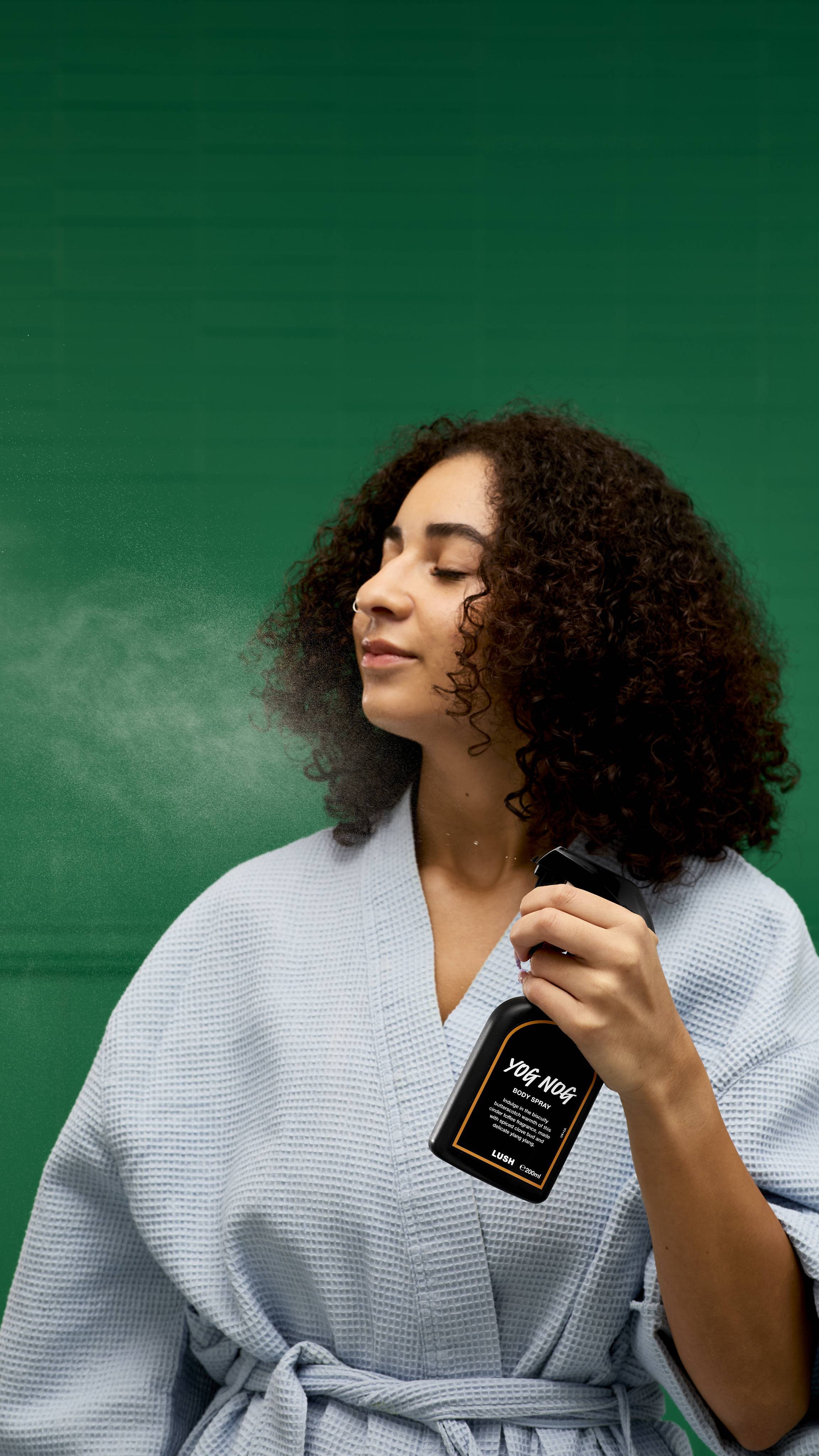 
A person with their eyes closed in bliss wears a light blue robe and holds a black LUSH "Yog Nog Body Spray" bottle. A fine mist of spray is visible on their face against a vibrant green background.