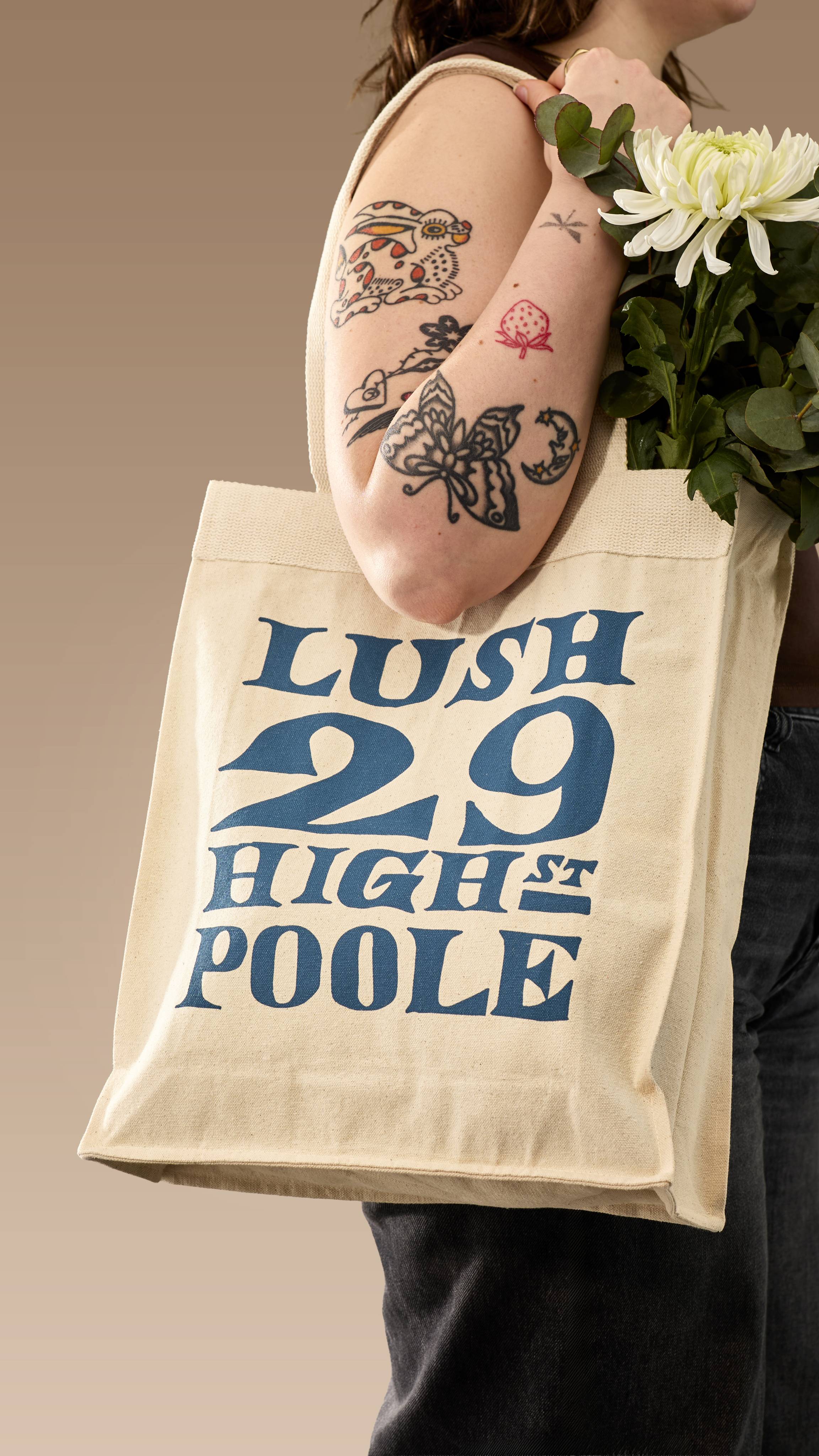 A model with several tattoos on their arm carries a cream canvas tote bag over their shoulder. The bag contains white flowers and features large navy-blue text reading ‘LUSH 29 HIGH ST POOLE’ in a vintage-style font.