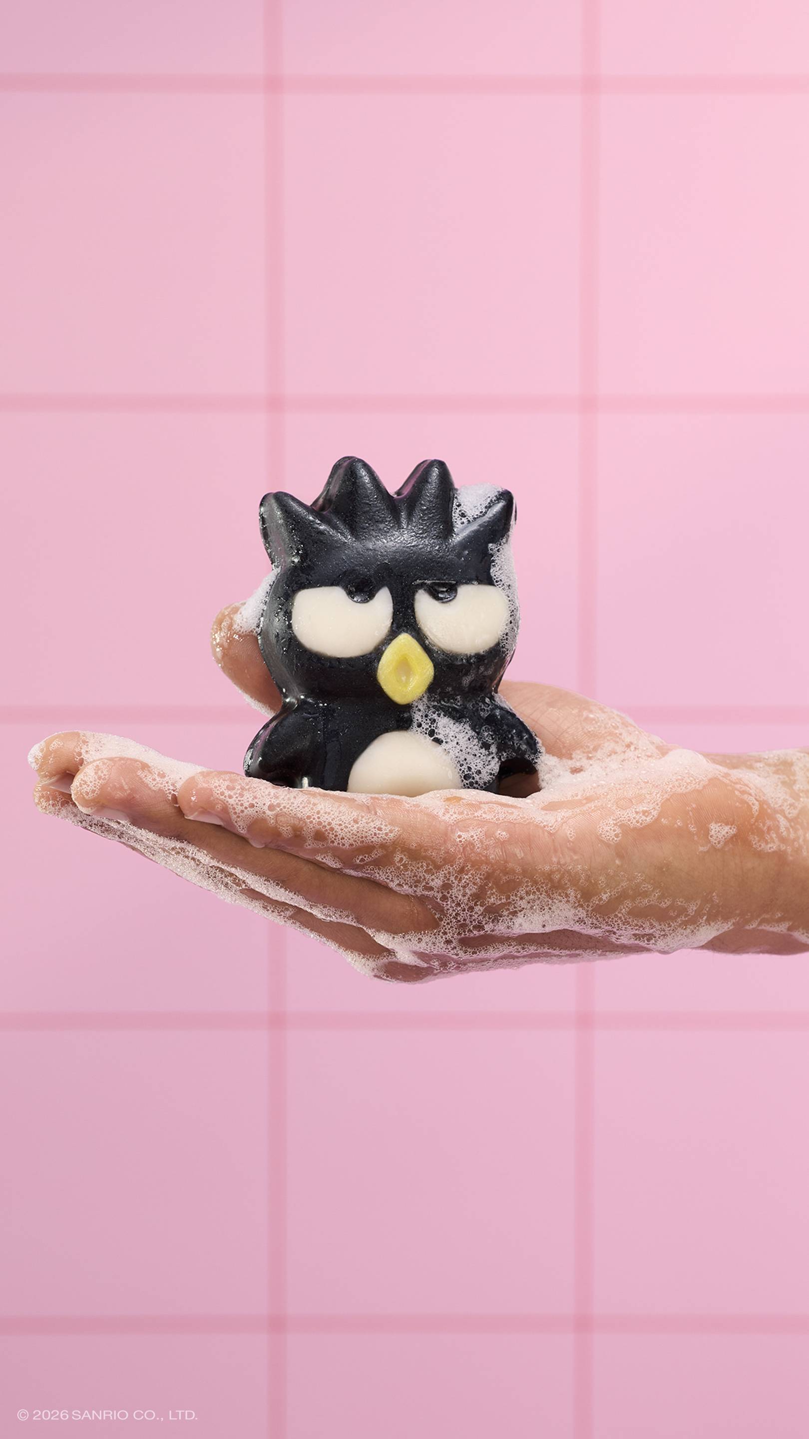 A soapy hand holds the black penguin-shaped "Bad Badtz-Maru" character soap, covered in white bubbles. The scene is set against a background of pink square tiles. 