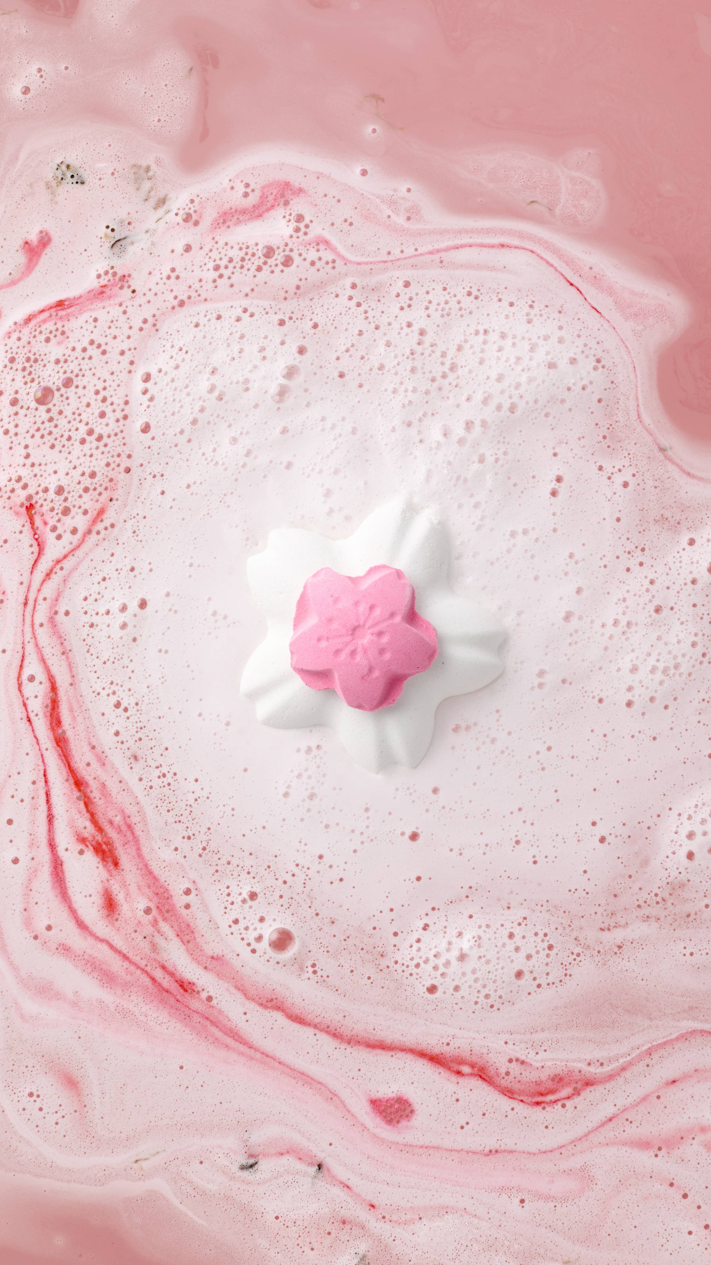 An aerial view of the "Blooming Cherry Blossoms" bath bomb with a smaller pink flower centre floats in the middle of a bath. It is surrounded by a large circle of frothy white bubbles and swirling patterns of vivid pink in the water.