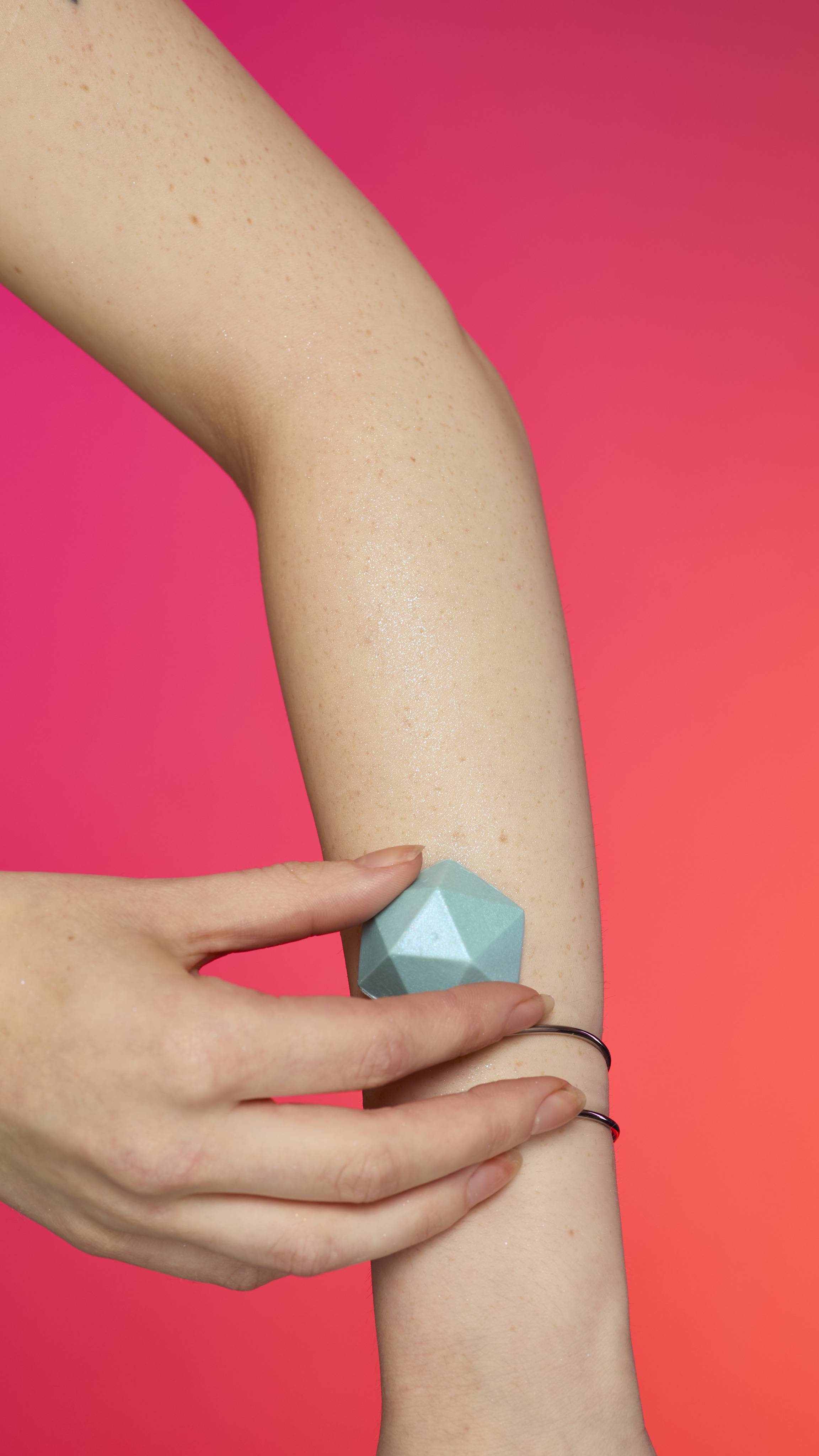 A hand with fair skin applies the faceted, pyramid-shaped Blue glitterbug body bar to an arm. The arm wears a thin bracelet and has a subtle sheen of plastic free glitter against a vibrant gradient of magenta, pink, and orange.