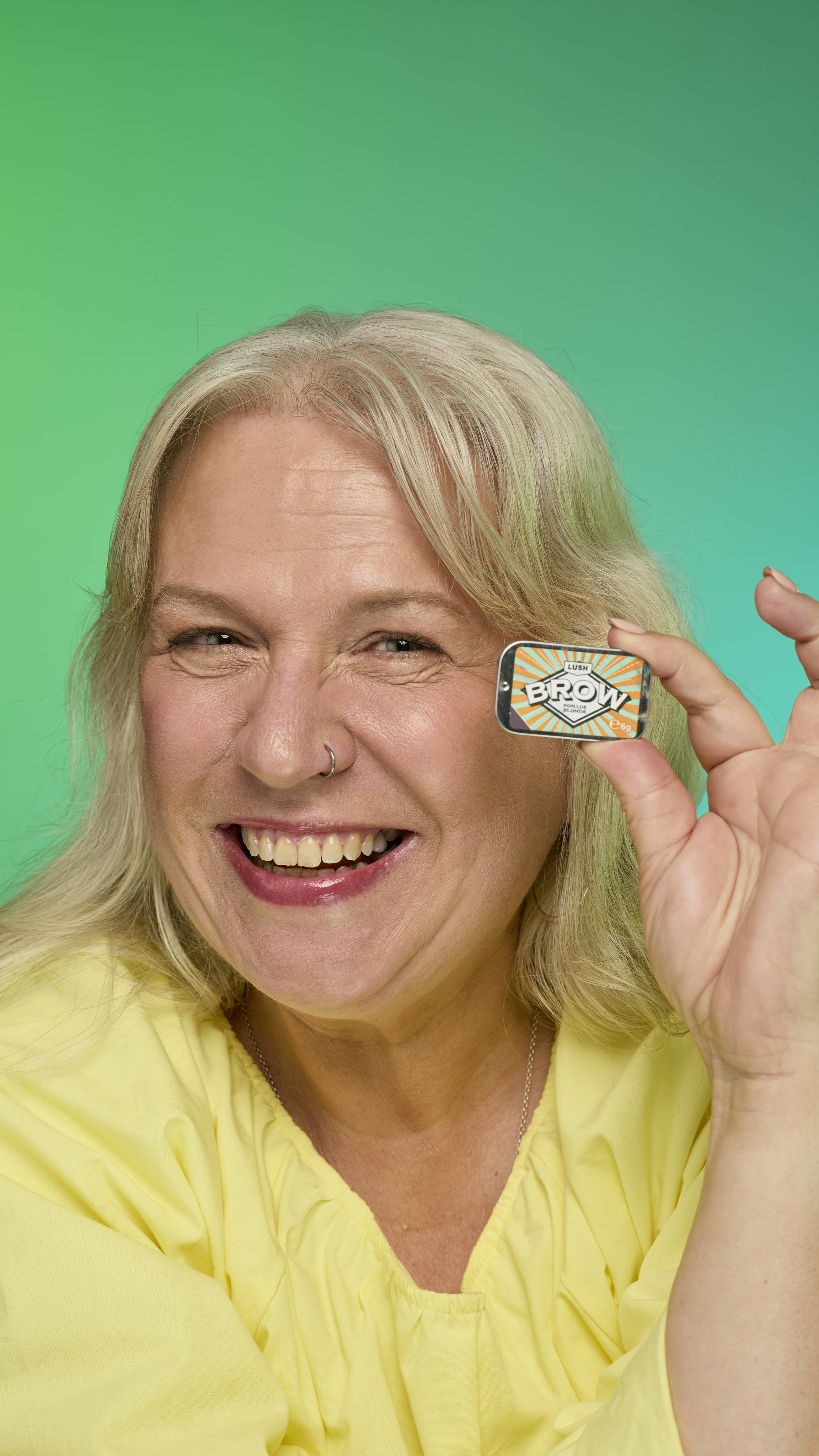 The image shows a model with blonde hair on a green background.  Their eyebrows look thick and full thanks to the Blonde brow pomade. They are holding up the metal tin of Blonde brow pomade product up to their face.