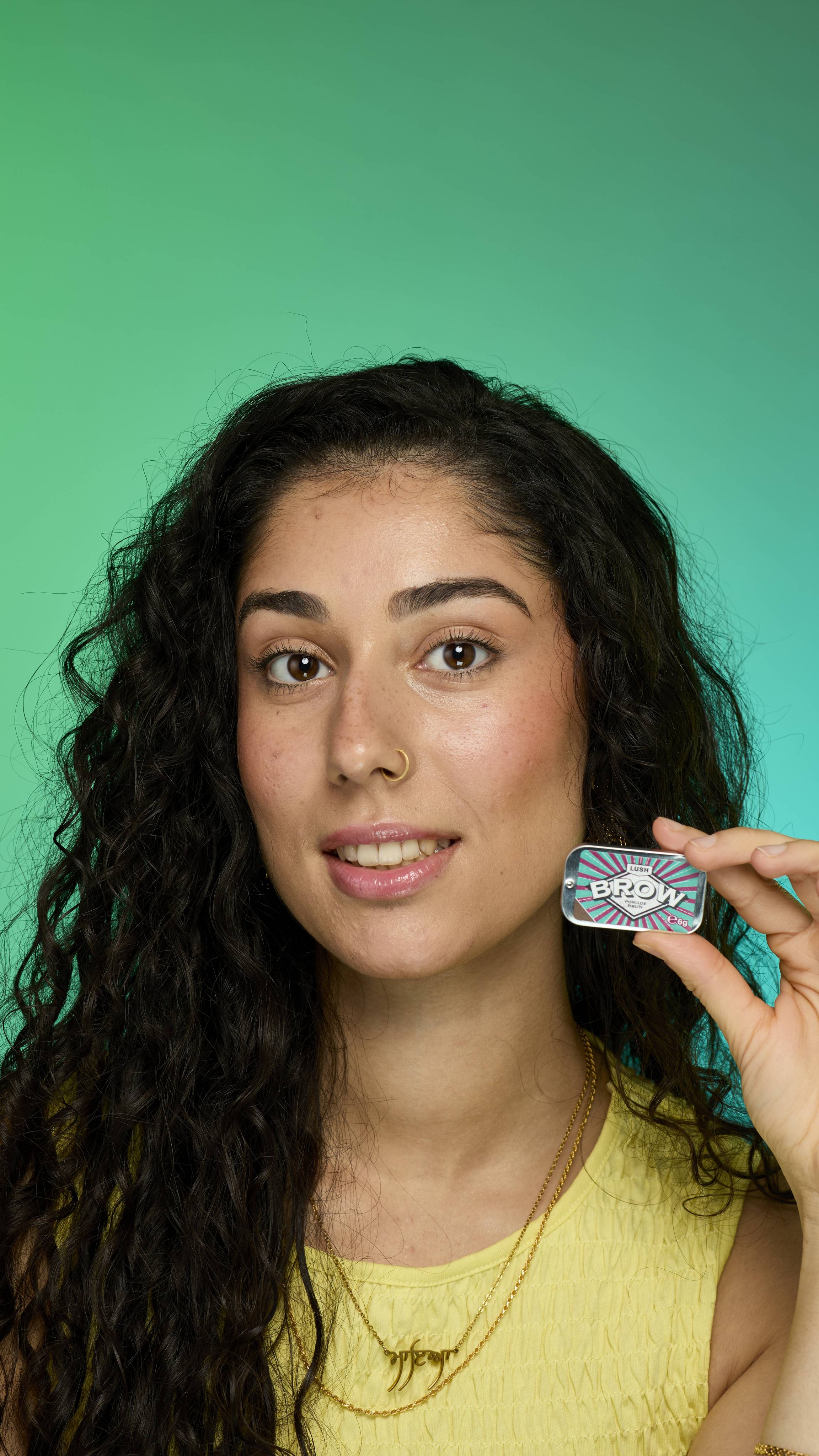 The image shows a model with dark curly hair on a green background.  Their eyebrows look thick and full thanks to the Brun brow pomade. They are holding up the metal tin of Brun brow pomade product up to their face.
