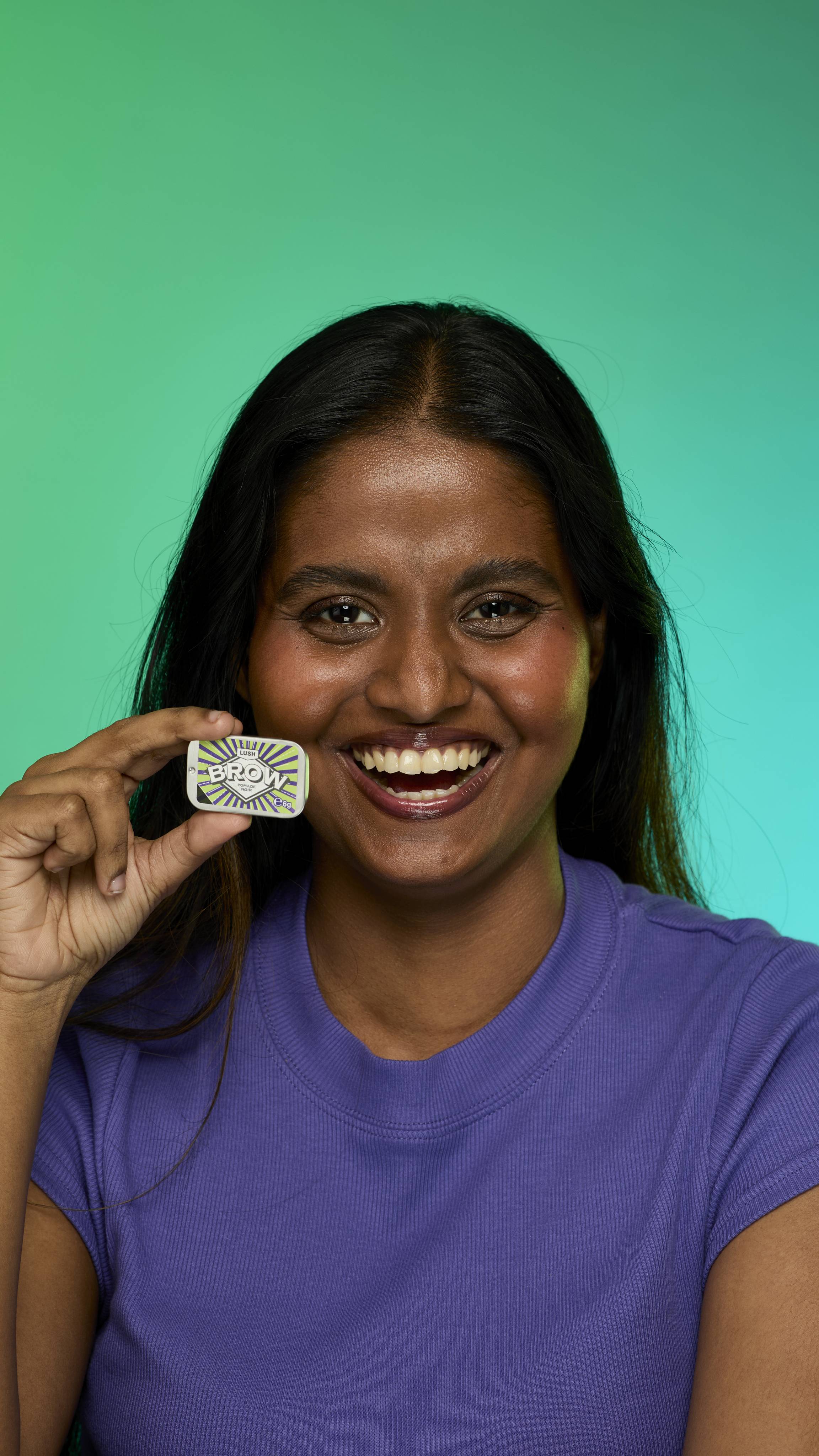The image shows a model with dark straight hair on a green background.  Their eyebrows look thick and full thanks to the Noir brow pomade. They are holding up the metal tin of Noir brow pomade product up to their face.