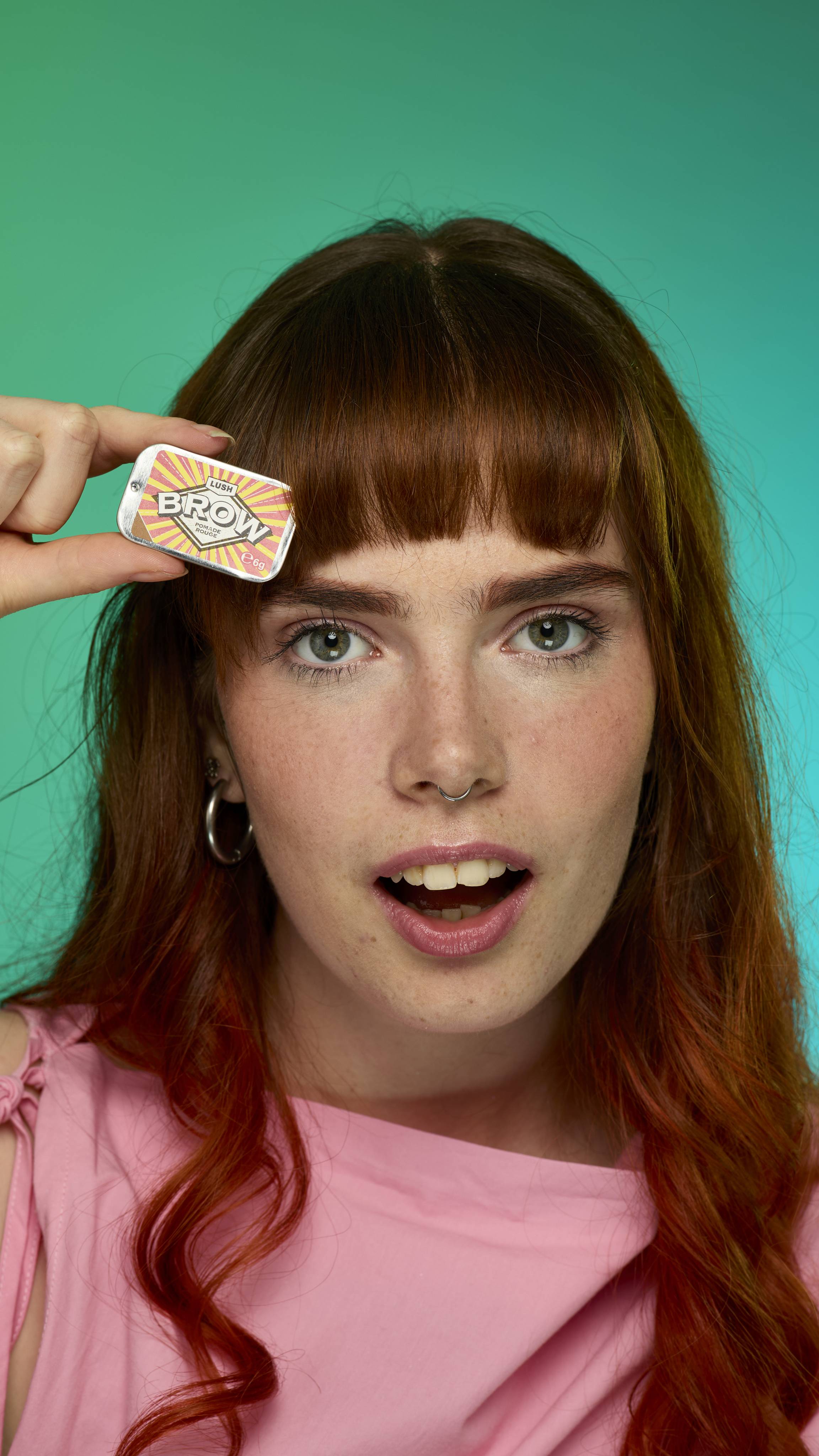 The image shows a model with fringed auburn hair on a green background.  Their eyebrows look thick and full thanks to the Rouge brow pomade. They are holding up the metal tin of Rouge brow pomade product up to their face.