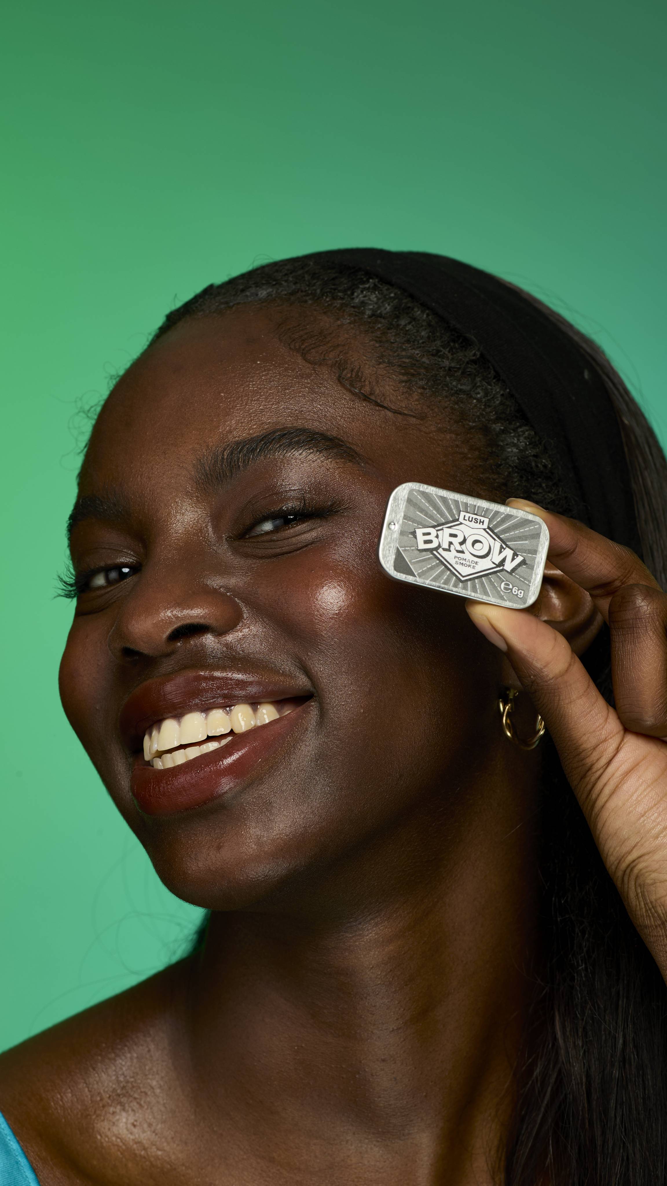 The image shows a model with dark straight hair on a green background.  Their eyebrows look thick and full thanks to the Smoke brow pomade. They are holding up the metal tin of Smoke brow pomade product up to their face.