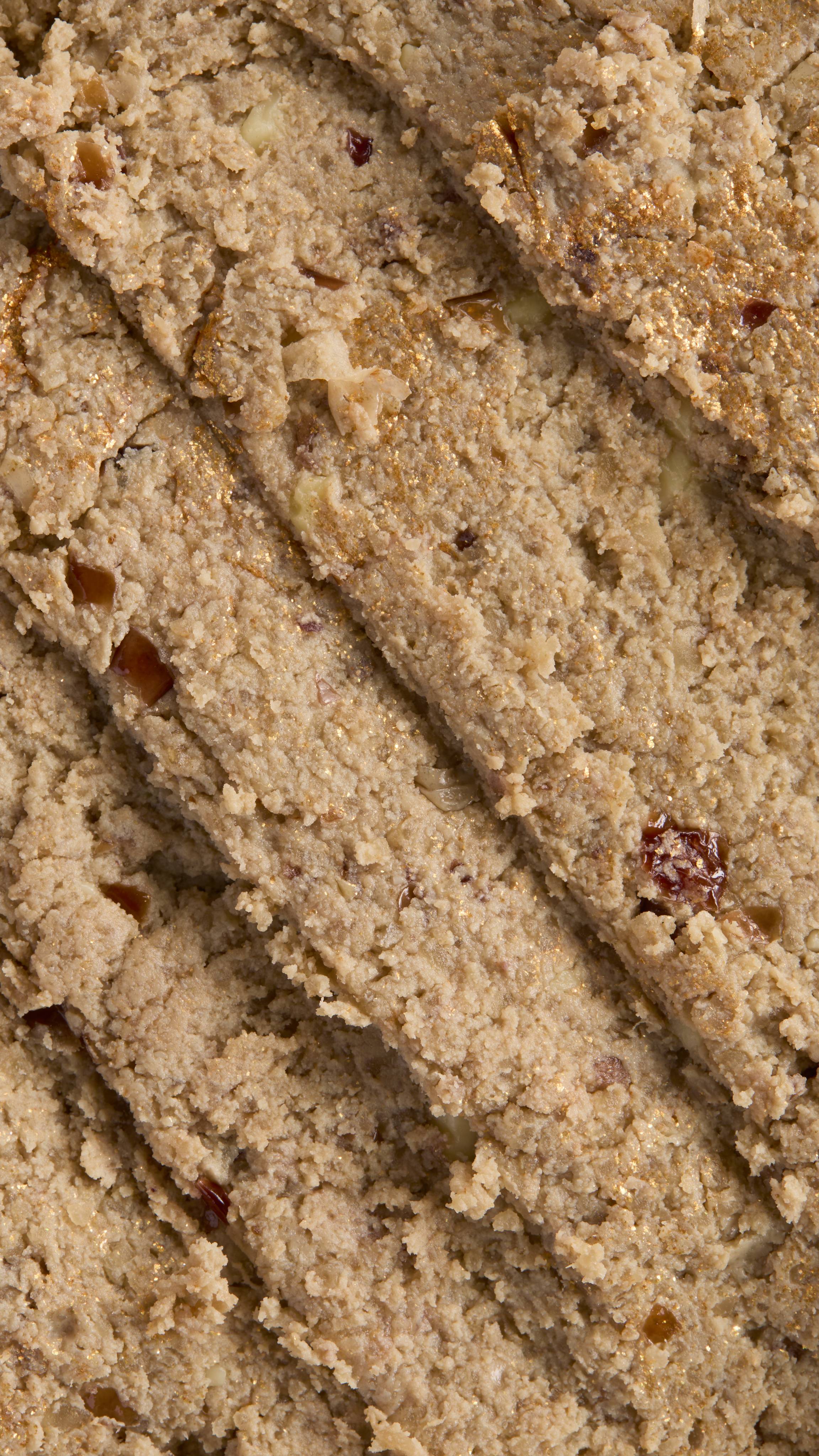 A close-up of a LUSH Bûche de Noël cleanser, a solid, light beige product with visible nut and cranberry pieces. It has a chunky texture and gold shimmer. The product has been sliced diagonally.
