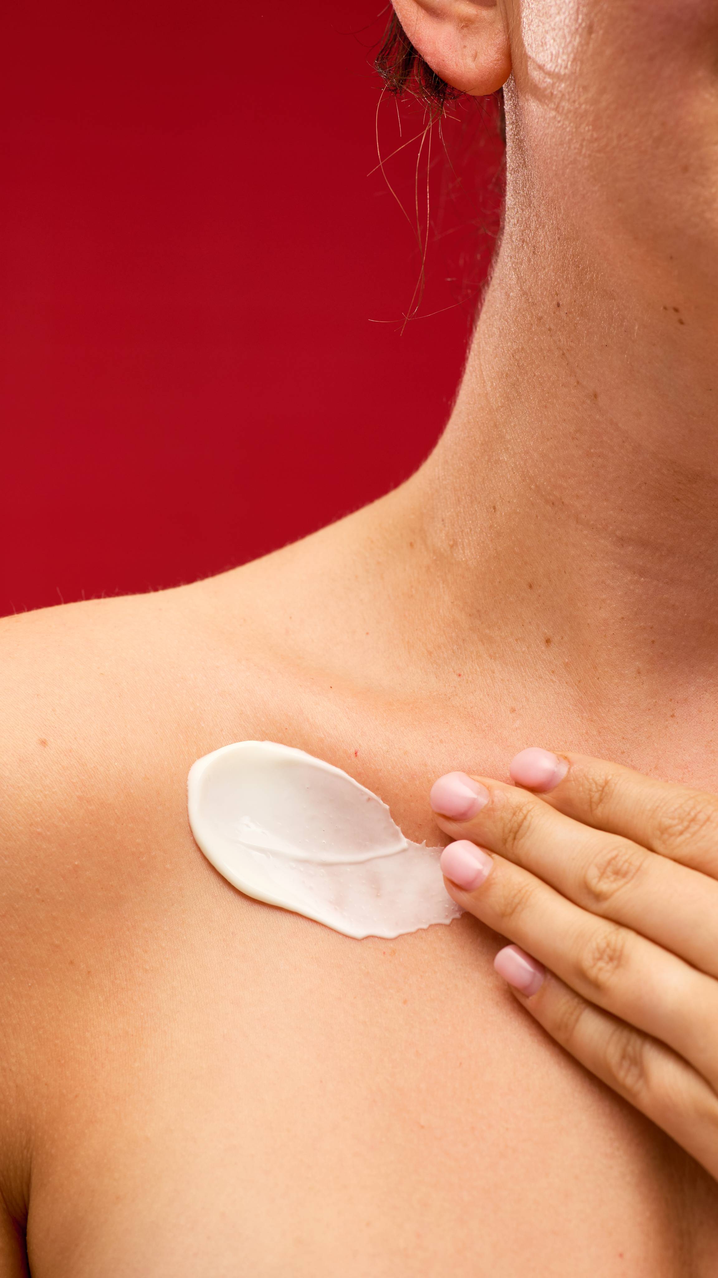 A close-up shot of a Celebrate body lotion swirled onto a person's collarbone. A hand with pink-painted nails is gently poised to massage it in, set against a vibrant red background.