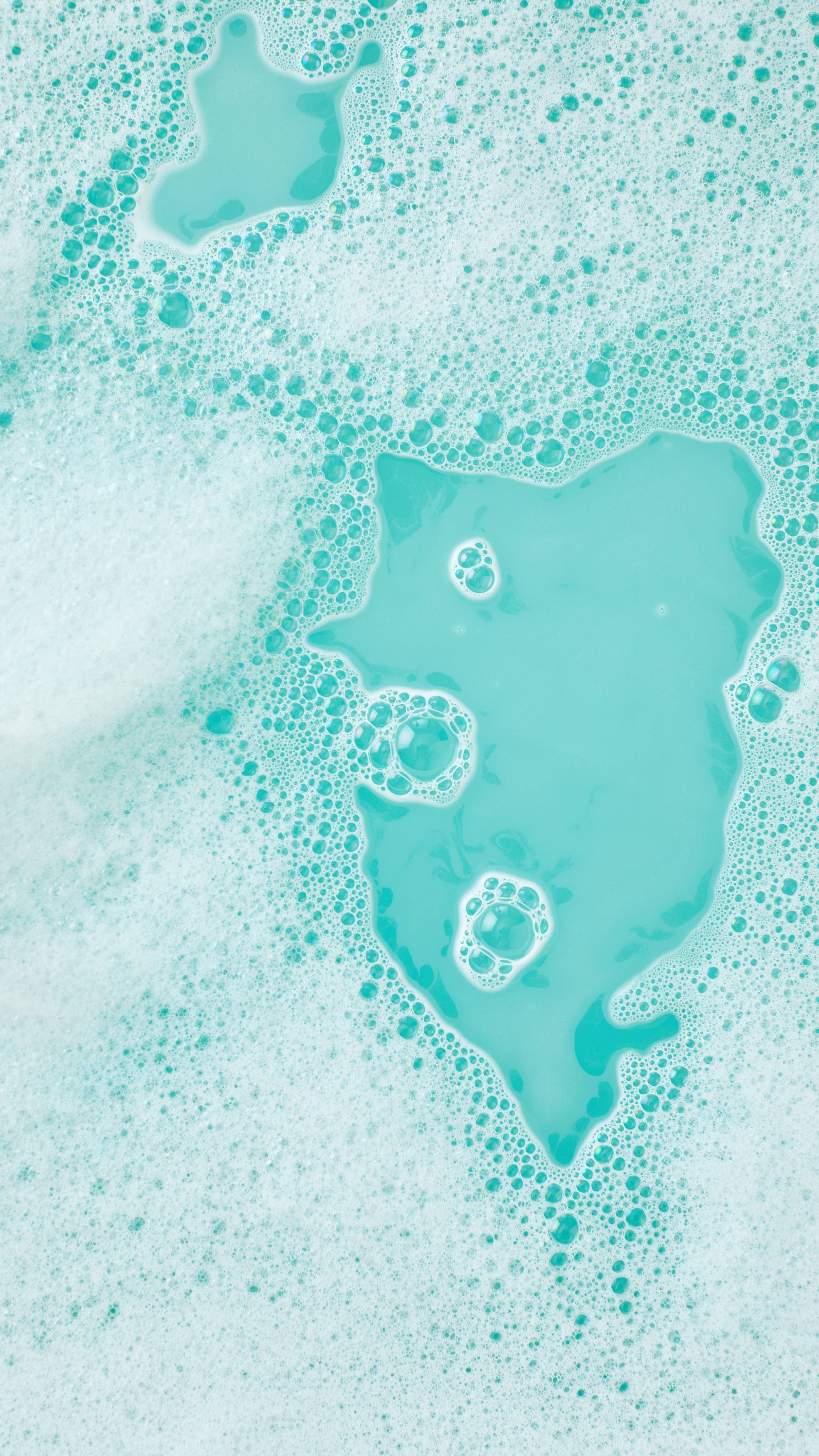 A close-up, aerial image of the bath water after using the Lush, Christmas Lights Fun product. There is a blanket of thick, fat bubbles with bright, teal-coloured bath water visible through a large gap.