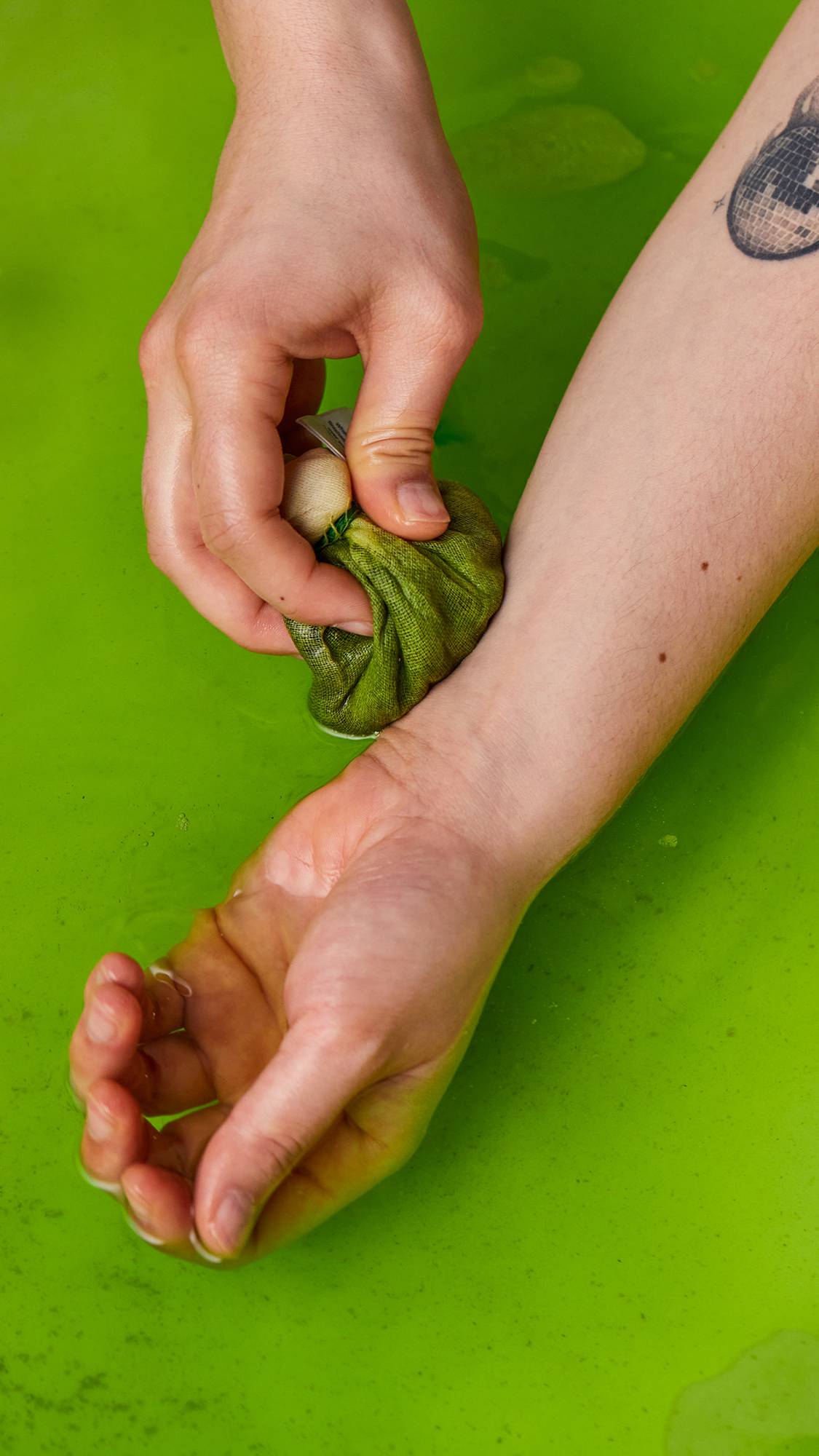 An aerial image of the bath showing the bright green water from the muslin-wrapped Energising Matcha And Peppermint Cataplasm bath bomb. Two arms reach in as gently soothe the remnants of the muslin pouch against their forearm.