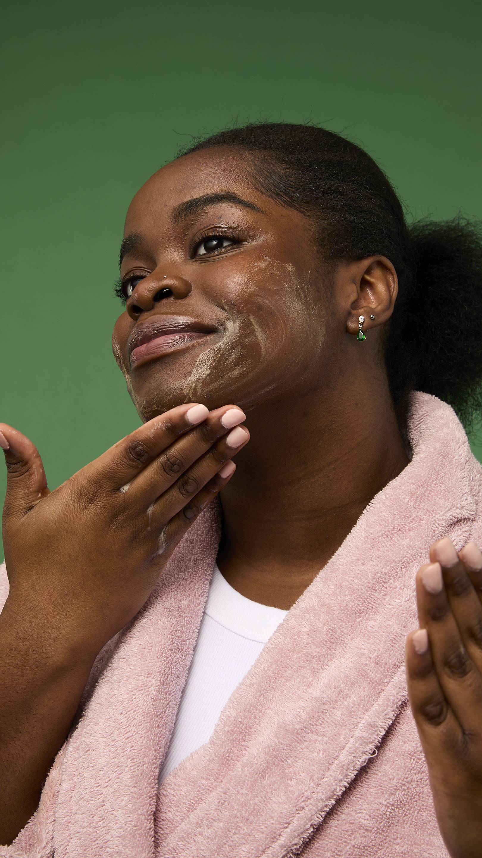 The image shows a model with dark skin, wearing a pink bathrobe from the chest up against a solid green background. They are rubbing golden-yellow suds from the Gingerbread gummy facial wash over their cheek.