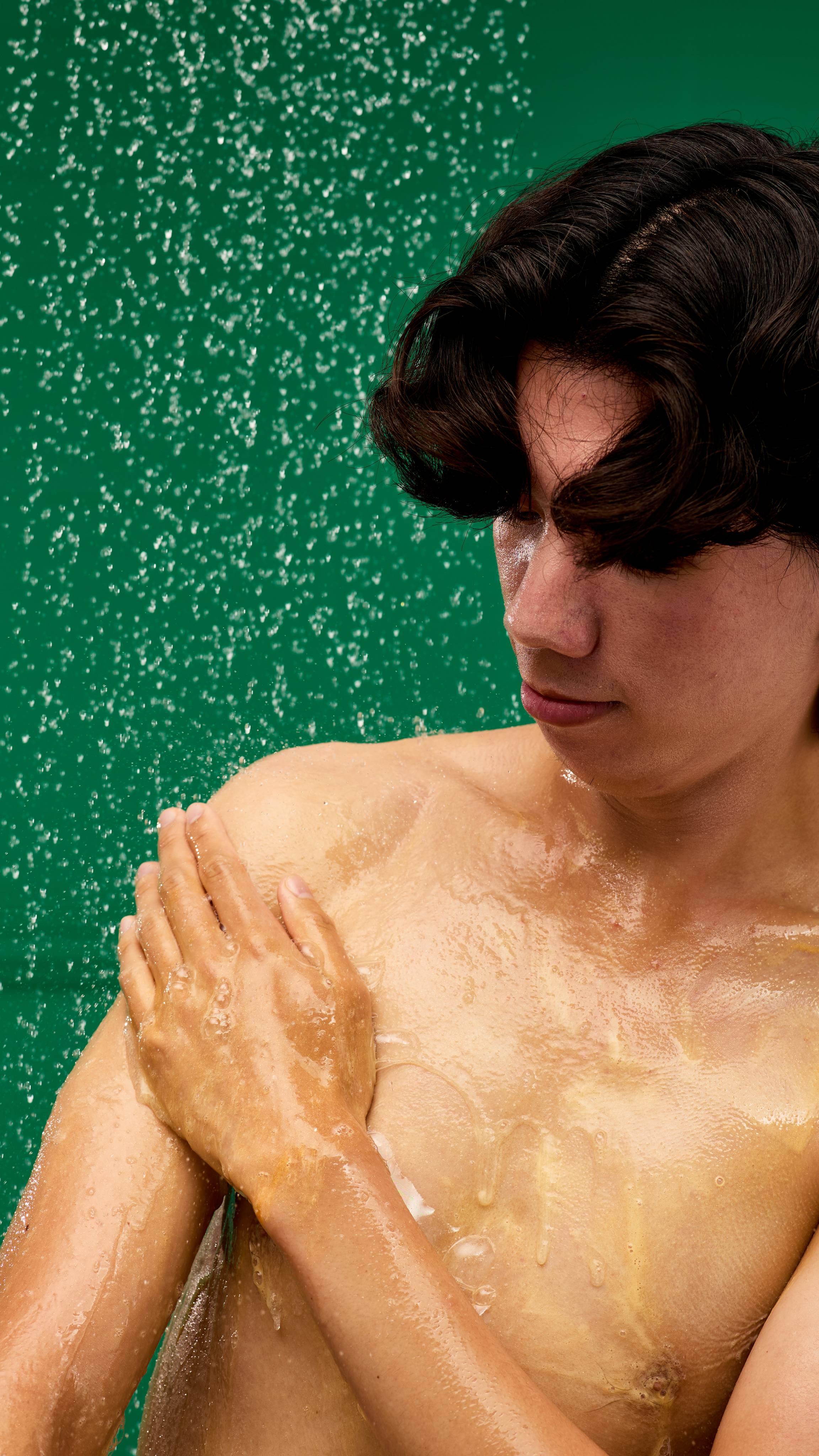 A person with wet hair lathers their chest and shoulder with LUSH Golden Pear shower gel. The glistening golden-orange product leaves a foamy residue, and water droplets are visible in the air against a vibrant green background.