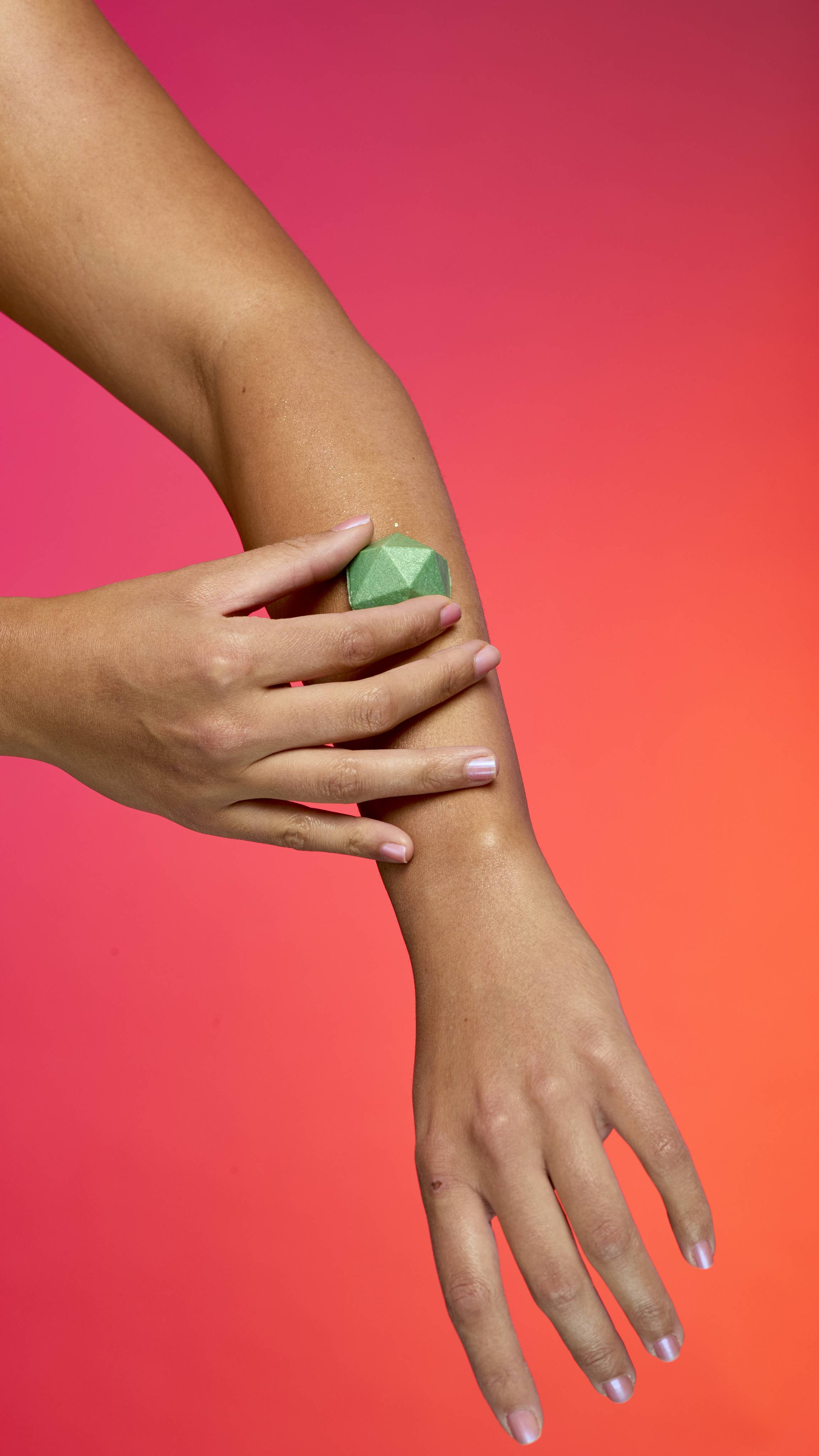 Two hands with tan skin apply the faceted, pyramid-shaped Green Glitterbug body bar to an arm. The skin has a sheen of plastic free glitter against a vibrant gradient of magenta, pink, and orange.