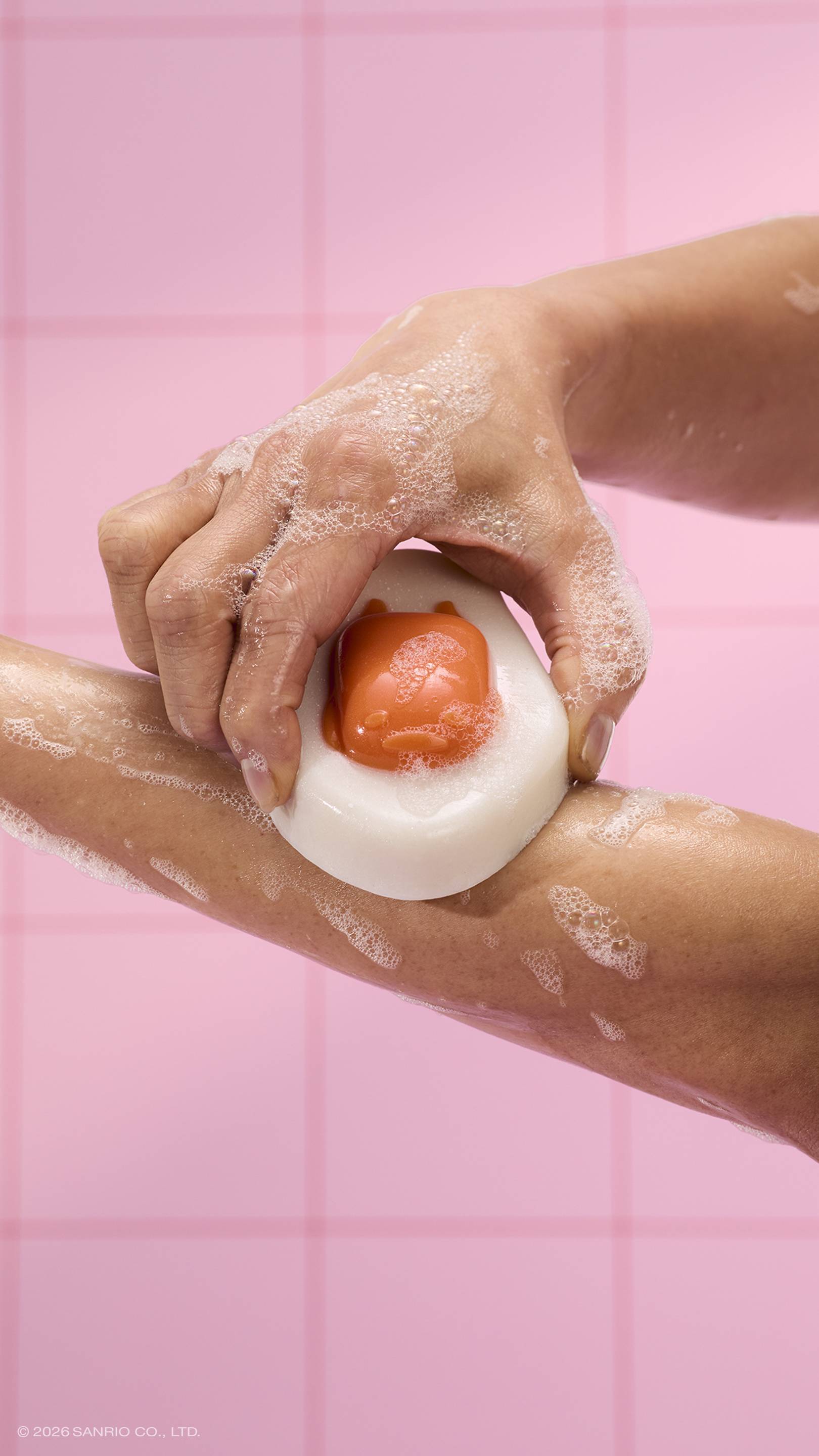 A close-up shows a person rubbing an egg-shaped "Gudetama" shower jelly against their arm. The glossy jelly features a sleepy orange face as the "yolk" and is covered in white bubbles, set against a pink tiled wall. 
