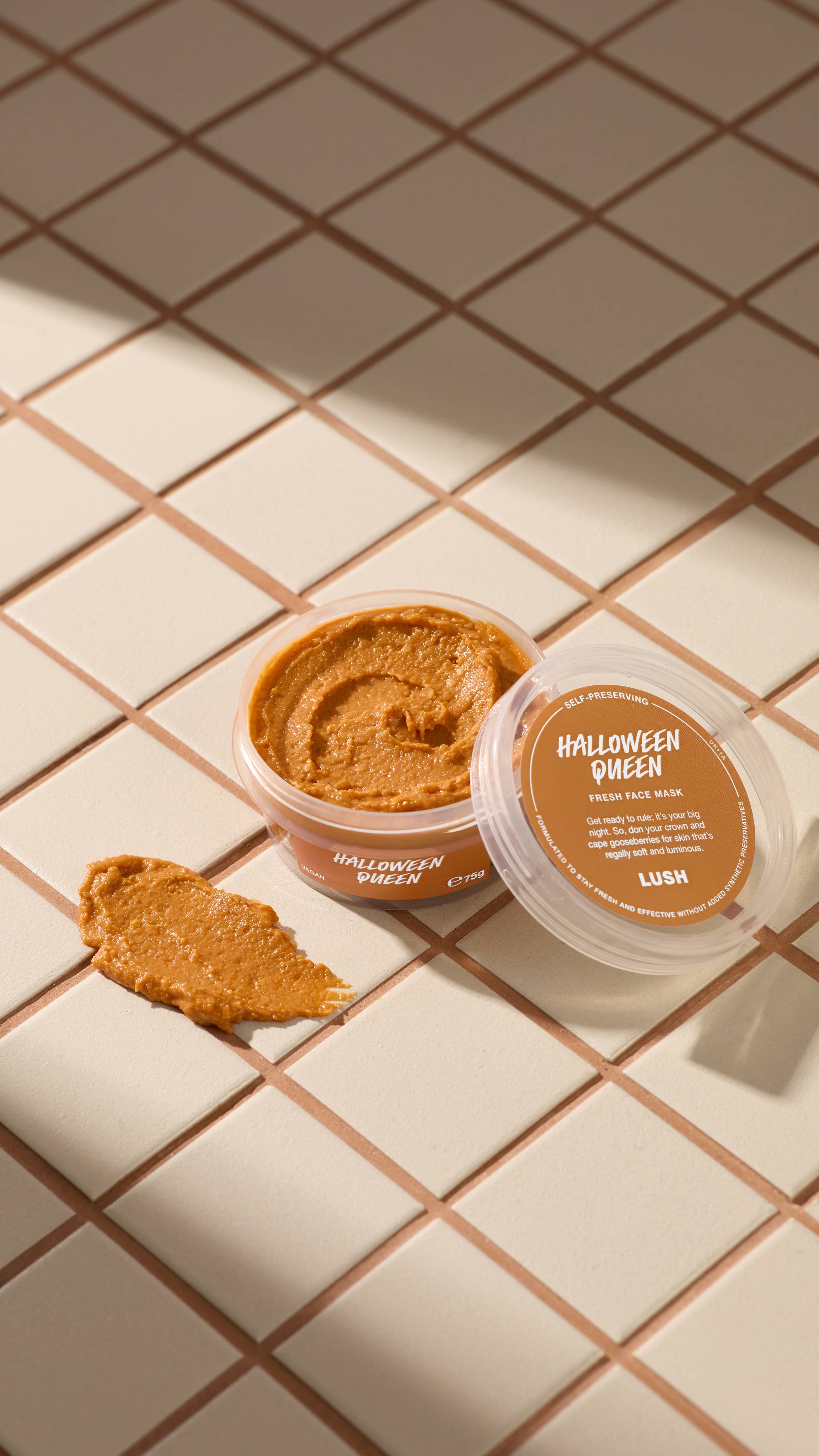 An open clear pot of Halloween Queen fresh face mask sits on beige tiles next to a textured orange smear of the product. The clear lid with an orange label rests against the pot. Sunny shadows cross the tiled surface.