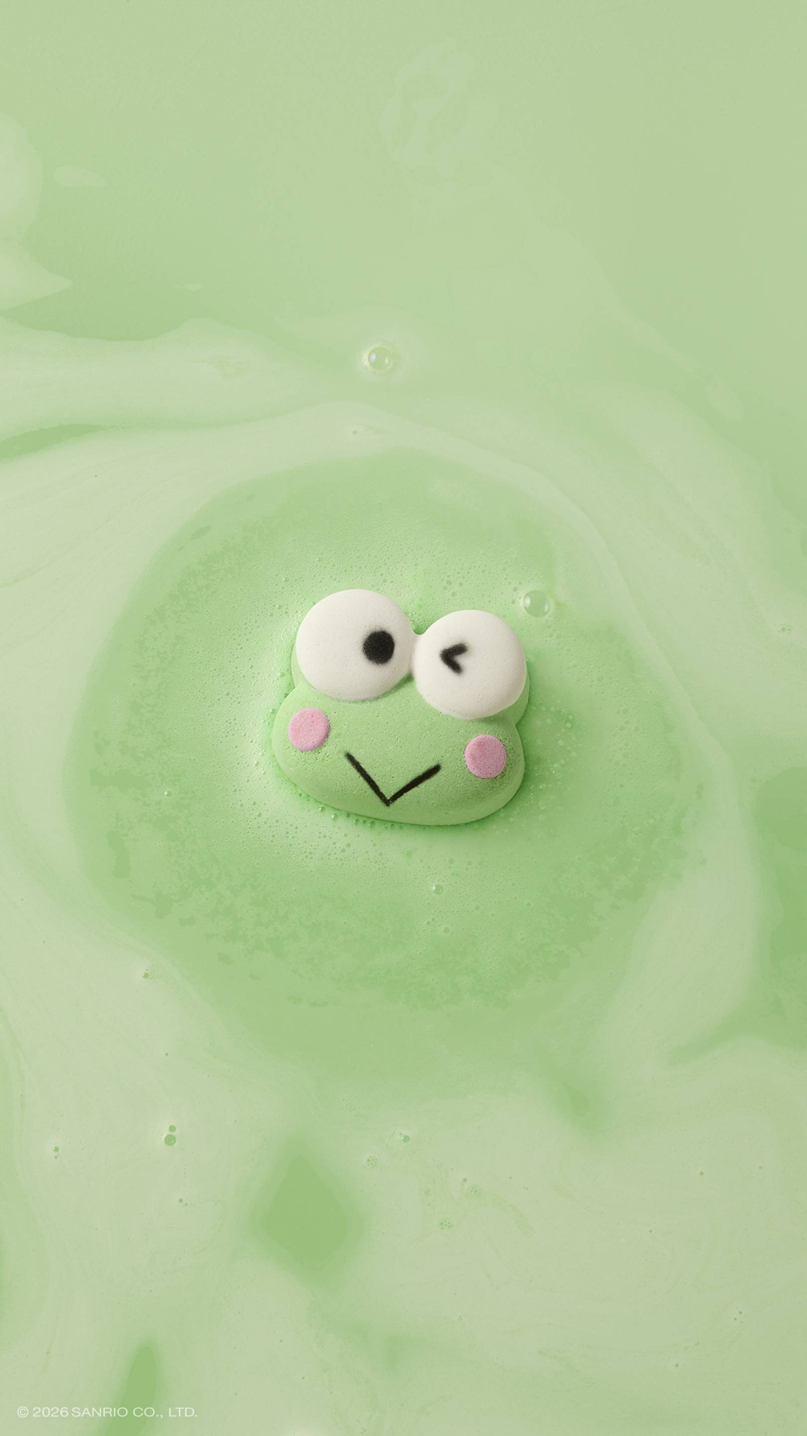 A green, winking frog-shaped bath bomb floats in a bath of lime-green water. It is surrounded by a soft circle of velvety foam as it dissolves, creating a creamy texture on the surface of the water.